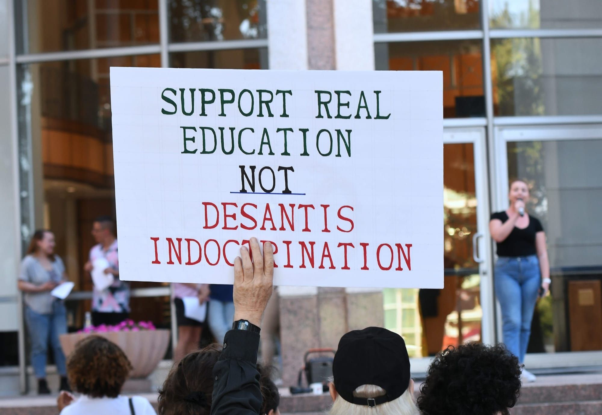 color photograph of an outdoor protest. a person holds up a large white sign with black text that reads "support real educati