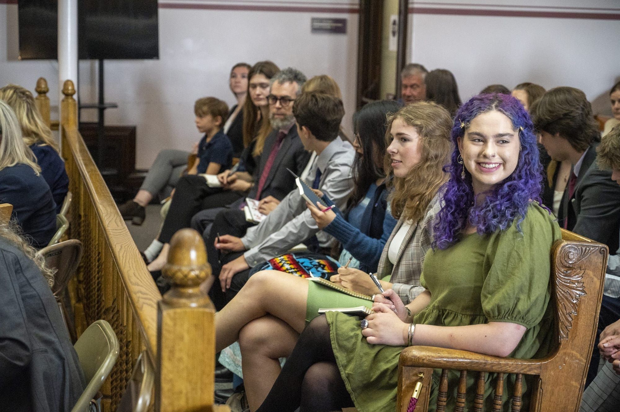 color photograph of a young person with curly purple hair just past their shoulders sitting on a courtroom bench. they're wea