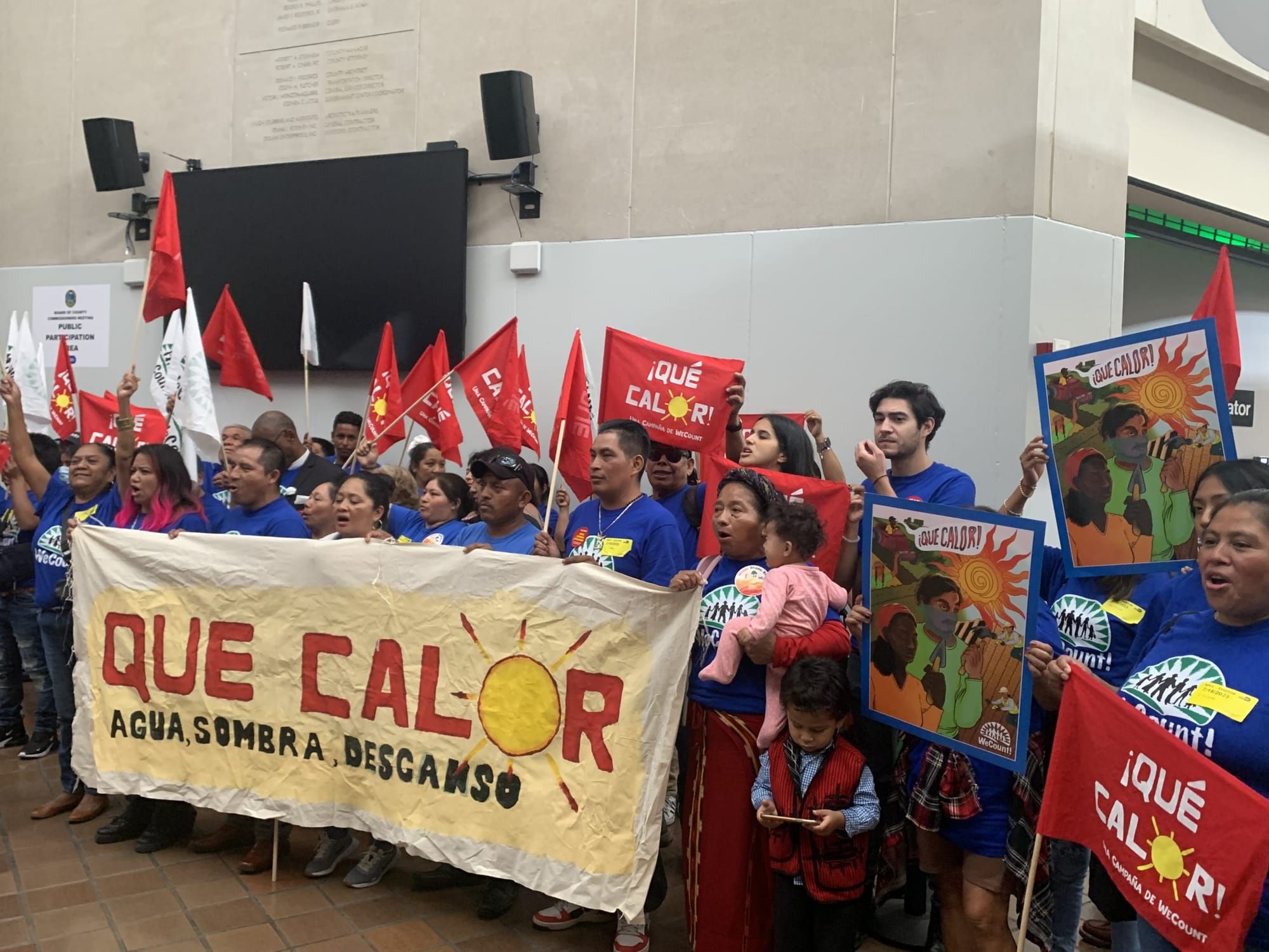 color photograph of an outdoor protest. people hold red cloth signs with white texts that reads "¡Que Calor!" and several peo