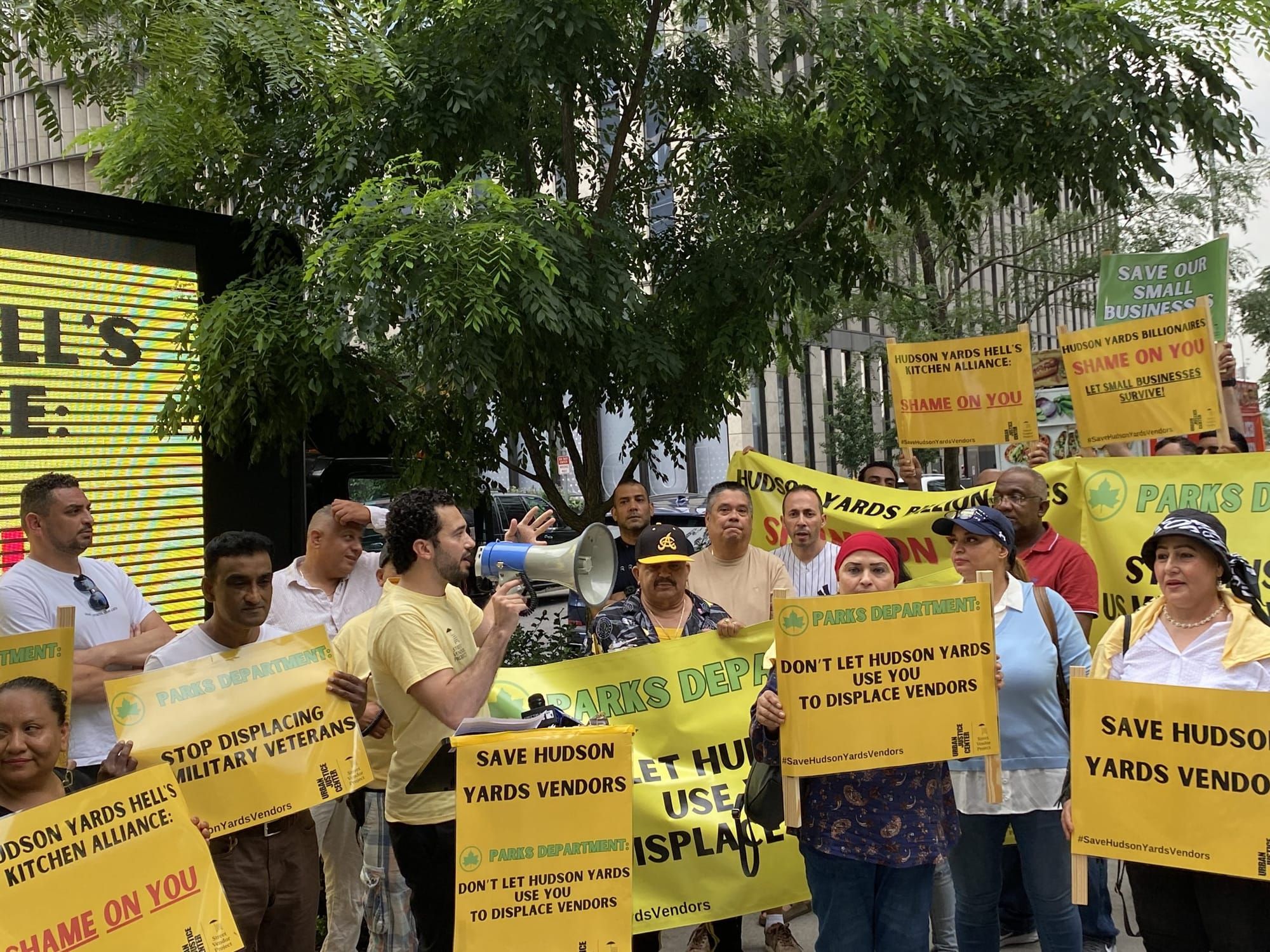 color photograph of an outdoor protest in support of new york street vendors. people hold up yellow signs with slogans such a