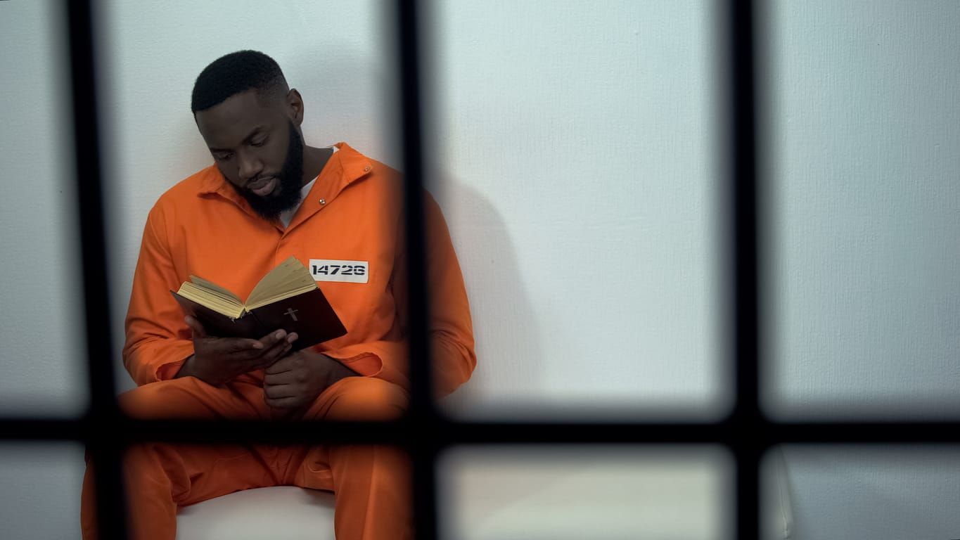 color photograph of a man in an orange prison jumpsuit sitting behind bars reading a book