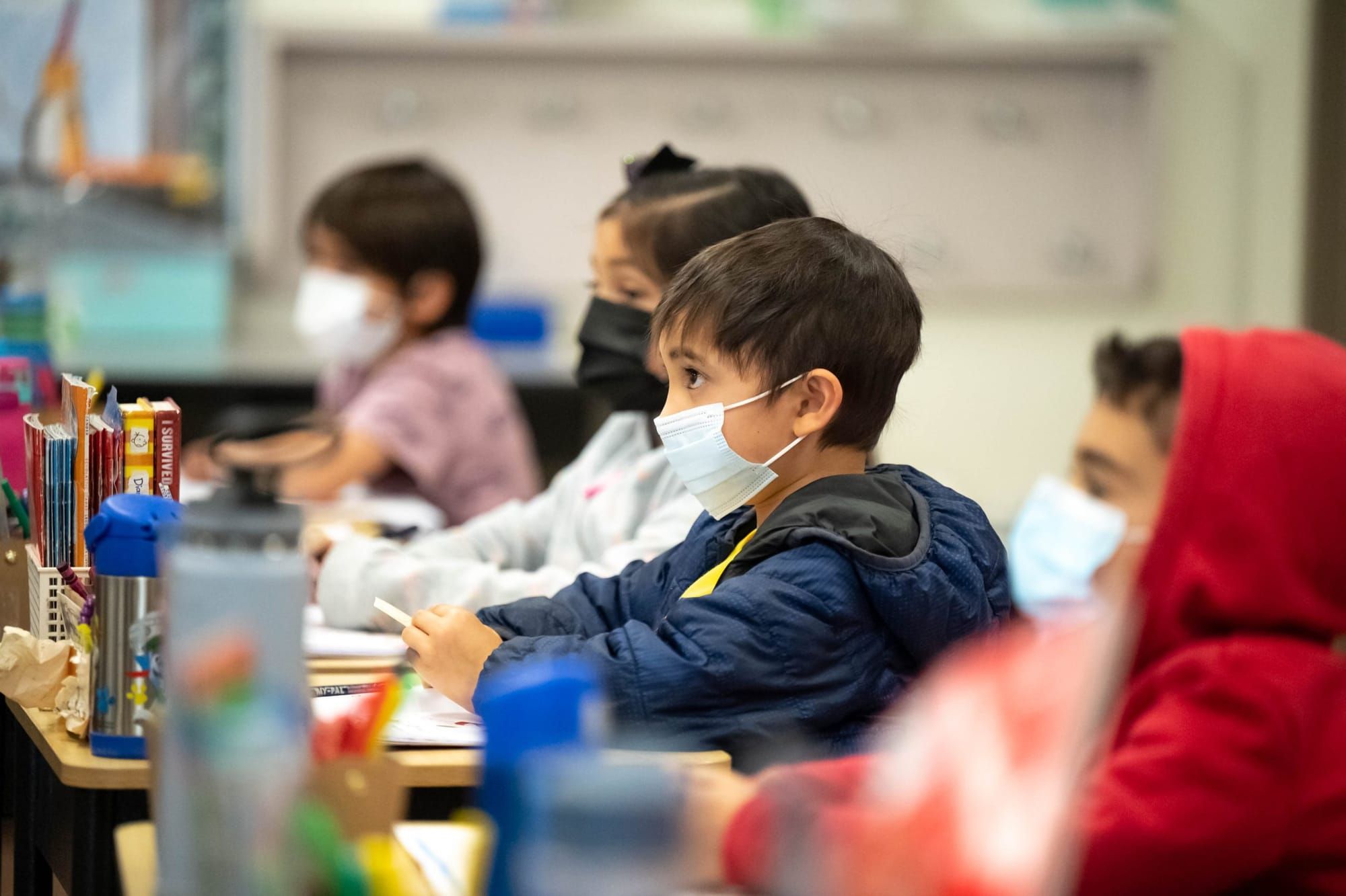 color photograph of the inside of a kindergarten classroom. four children sit in a diagonal at a table in the mid-ground. all