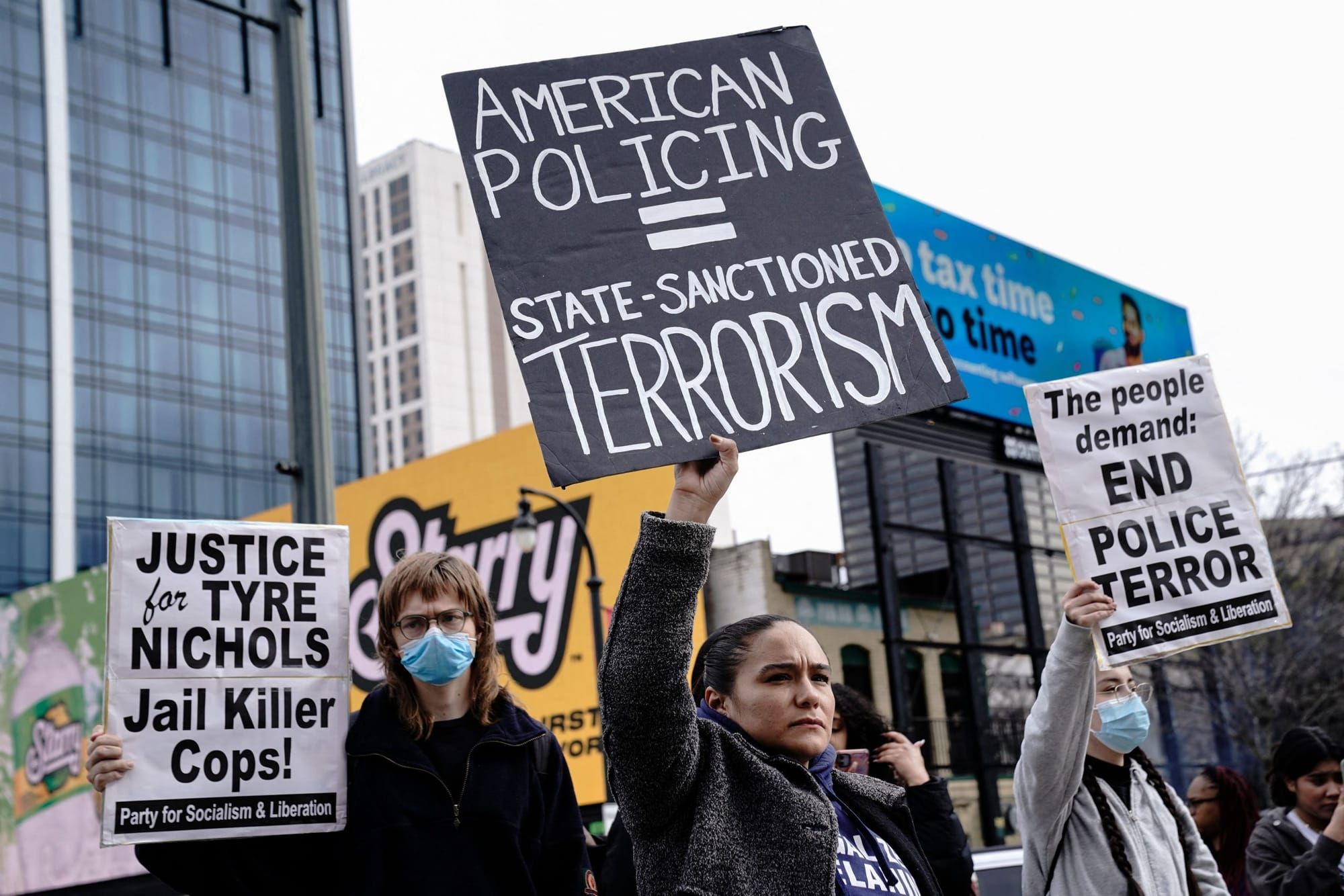 color photograph of an outdoor protest against police violence. a women in a dark grey fleece jacket holds up a black paper s