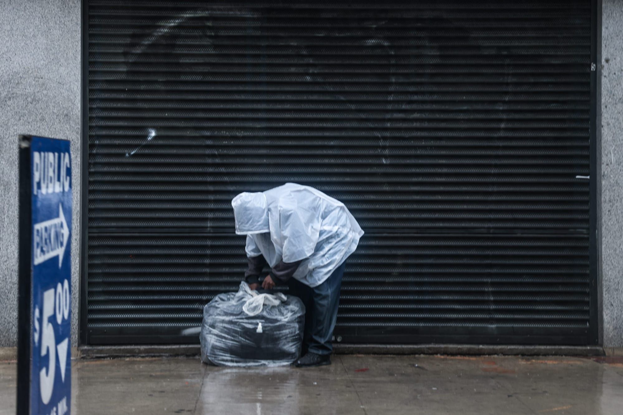 color photograph of a person wearing a plastic opaque white poncho bent over on a sidewalk rummaging through a plastic trash