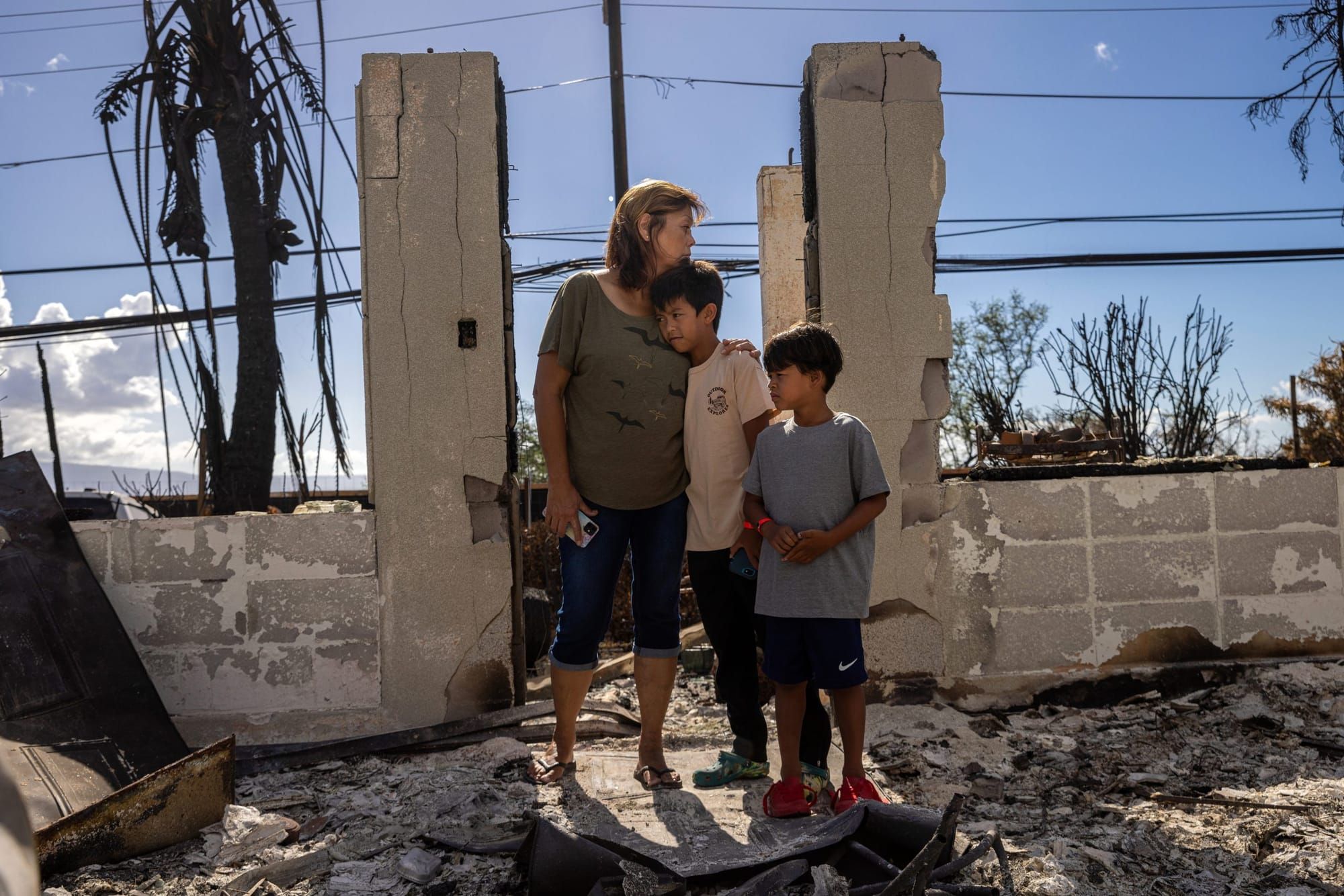 color photograph of a Hawaiian grandmother standing next to her two grandsons in front of the stone ruins of their house