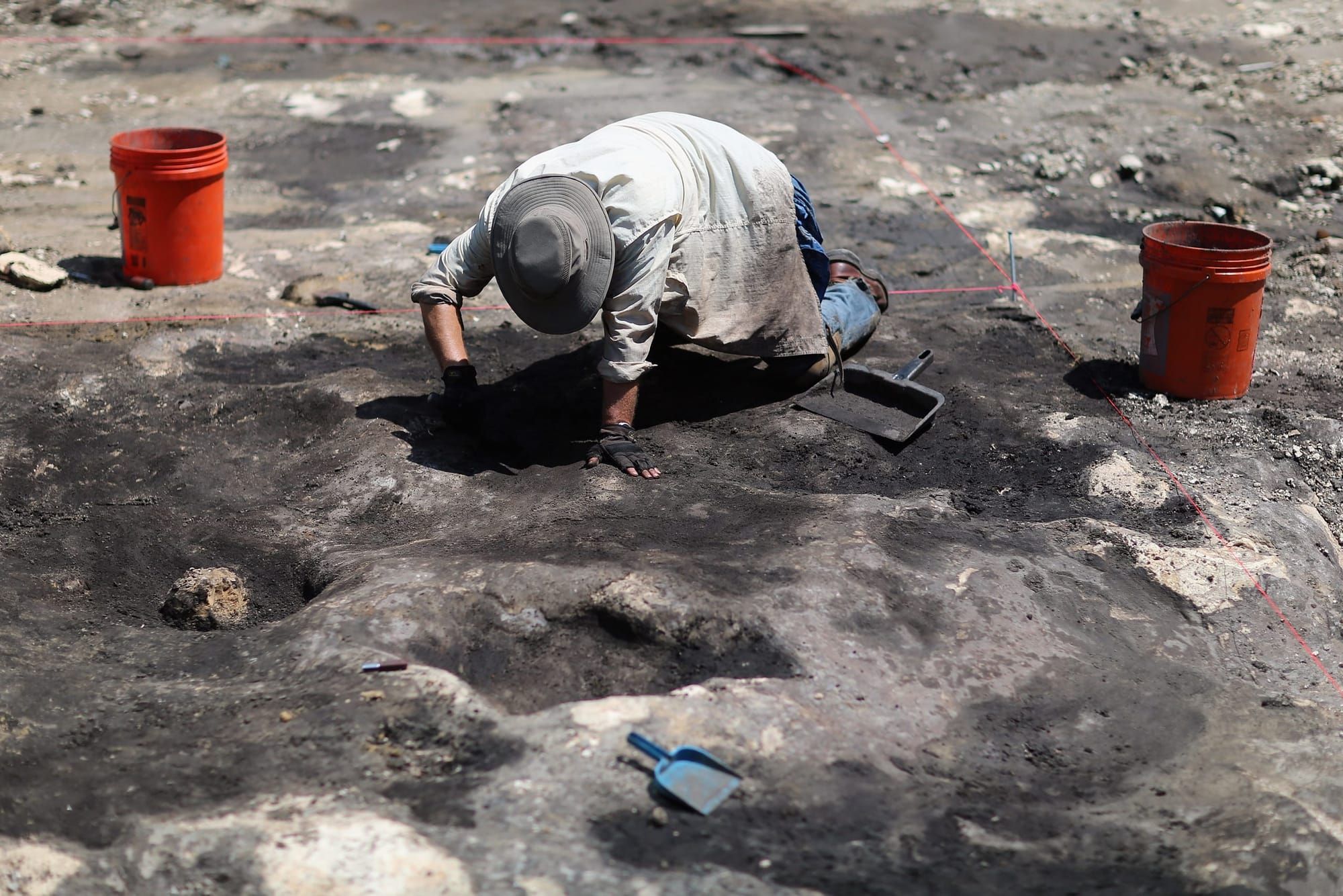 color photograph of an archaeologist on their hands and knees digging on a flat grey stone surface. red lines of string creat