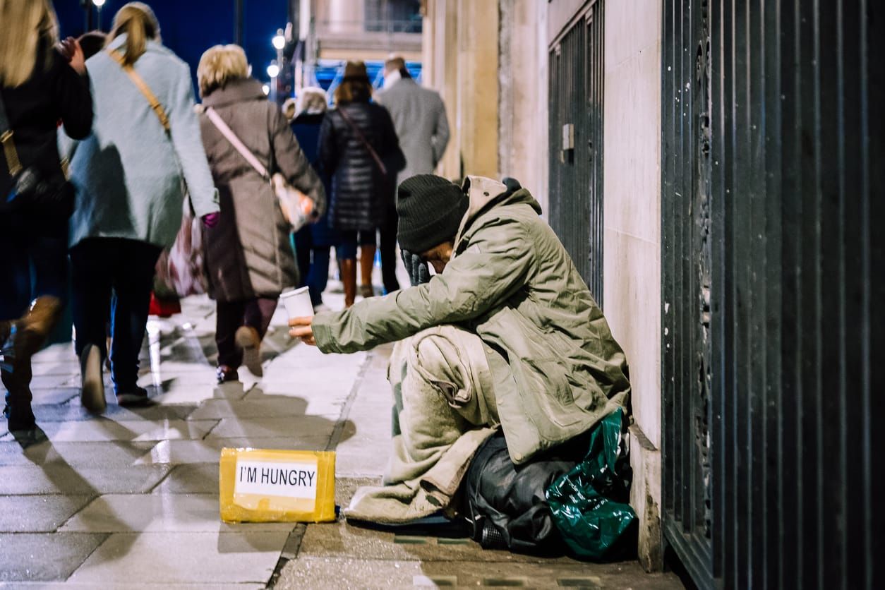 color photograph of a person wearing an oversized jacket and black beanie hat sitting on a sidewalk and holding out a cup as