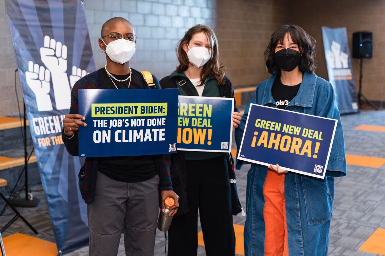 color photograph of three people standing inside wearing masks. they hold blue posters with printed text slogans supporting t