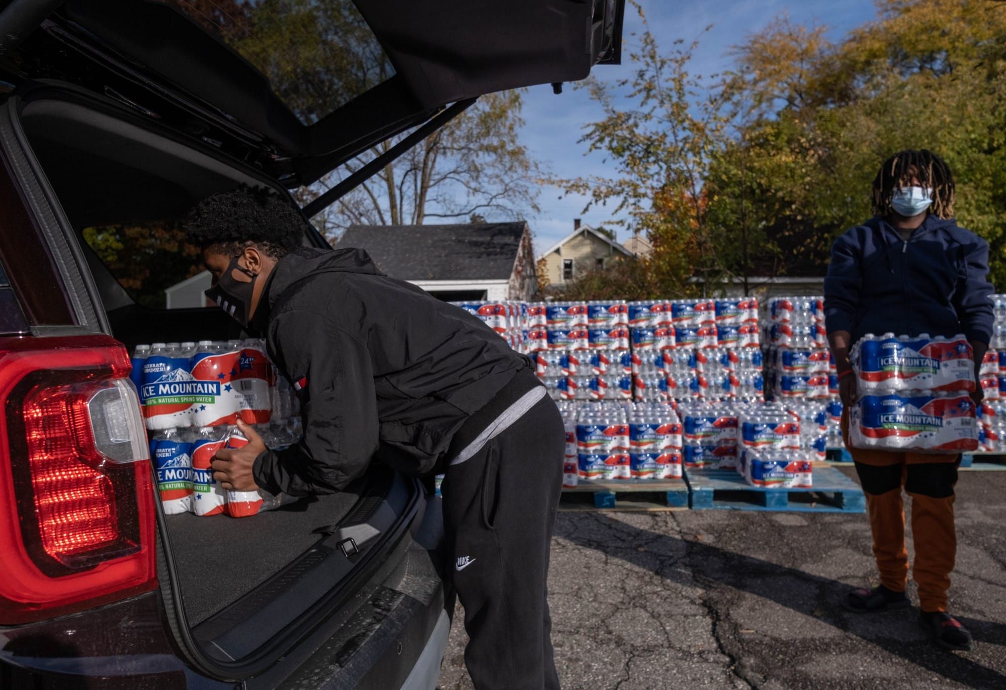 color photograph of two Black people in dark clothing loading packs of water into the back of an SUV car