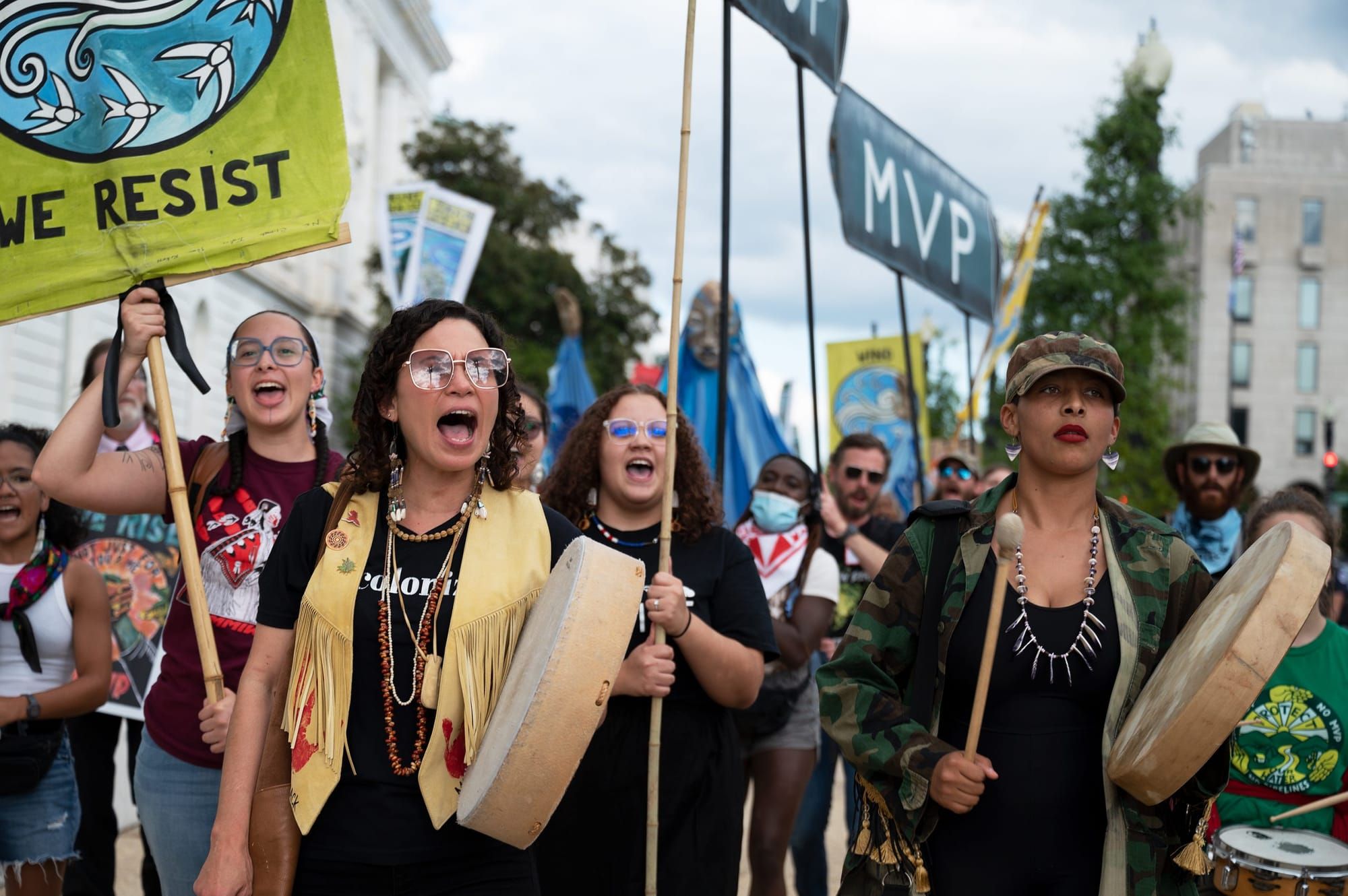 color photograph of an outdoor protest against the mountain valley pipeline. people in the foreground hold hand drums and peo