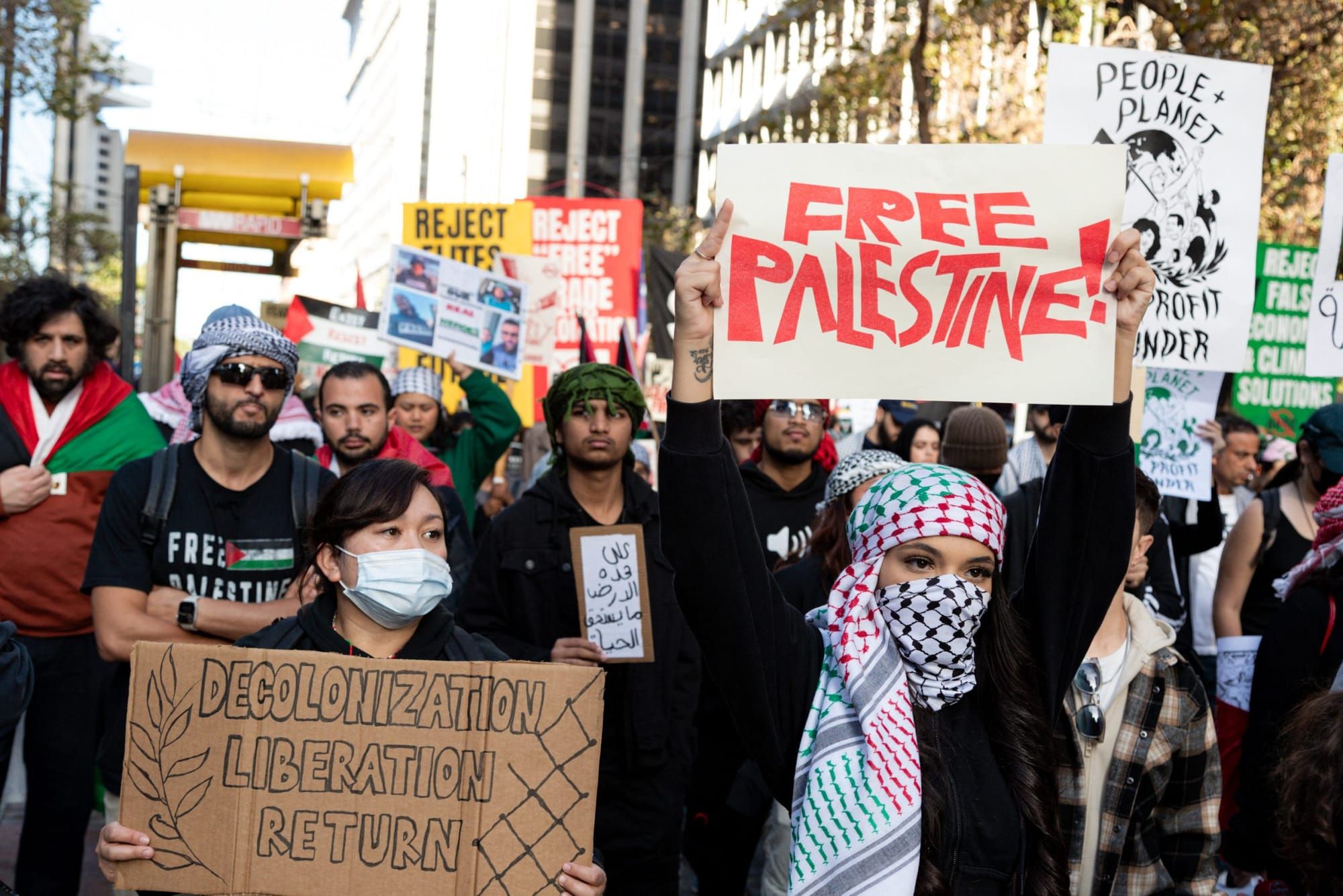 color photograph of an outdoor protest in support of palestine. people hold up paper and cardboard signs with handwritten mes