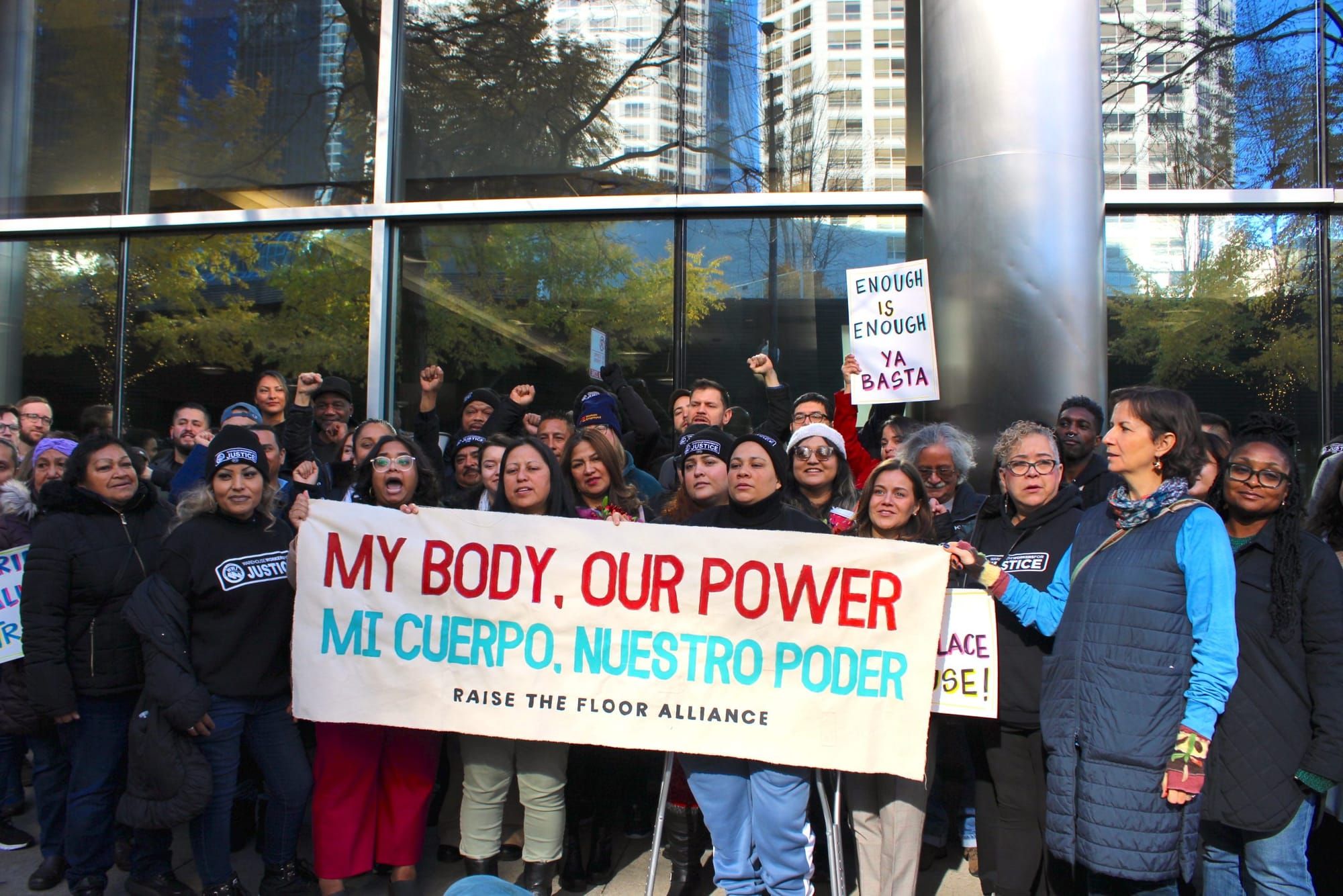 color photograph of an outdoor protest of mostly Latinx workers holding up a banner with red text that reads "My body, our po