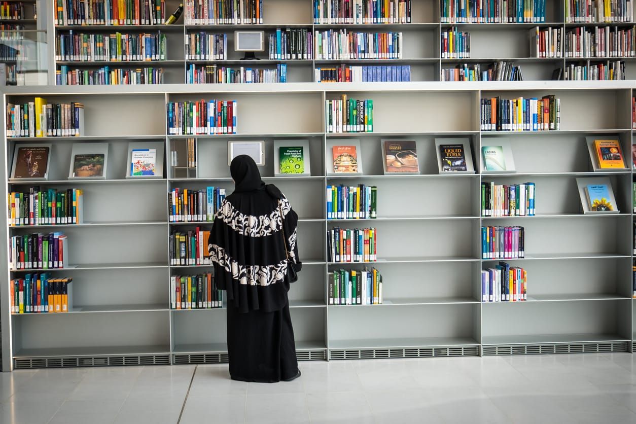 a woman black head covering and long black skirt stands in front of mostly empty bookshelves