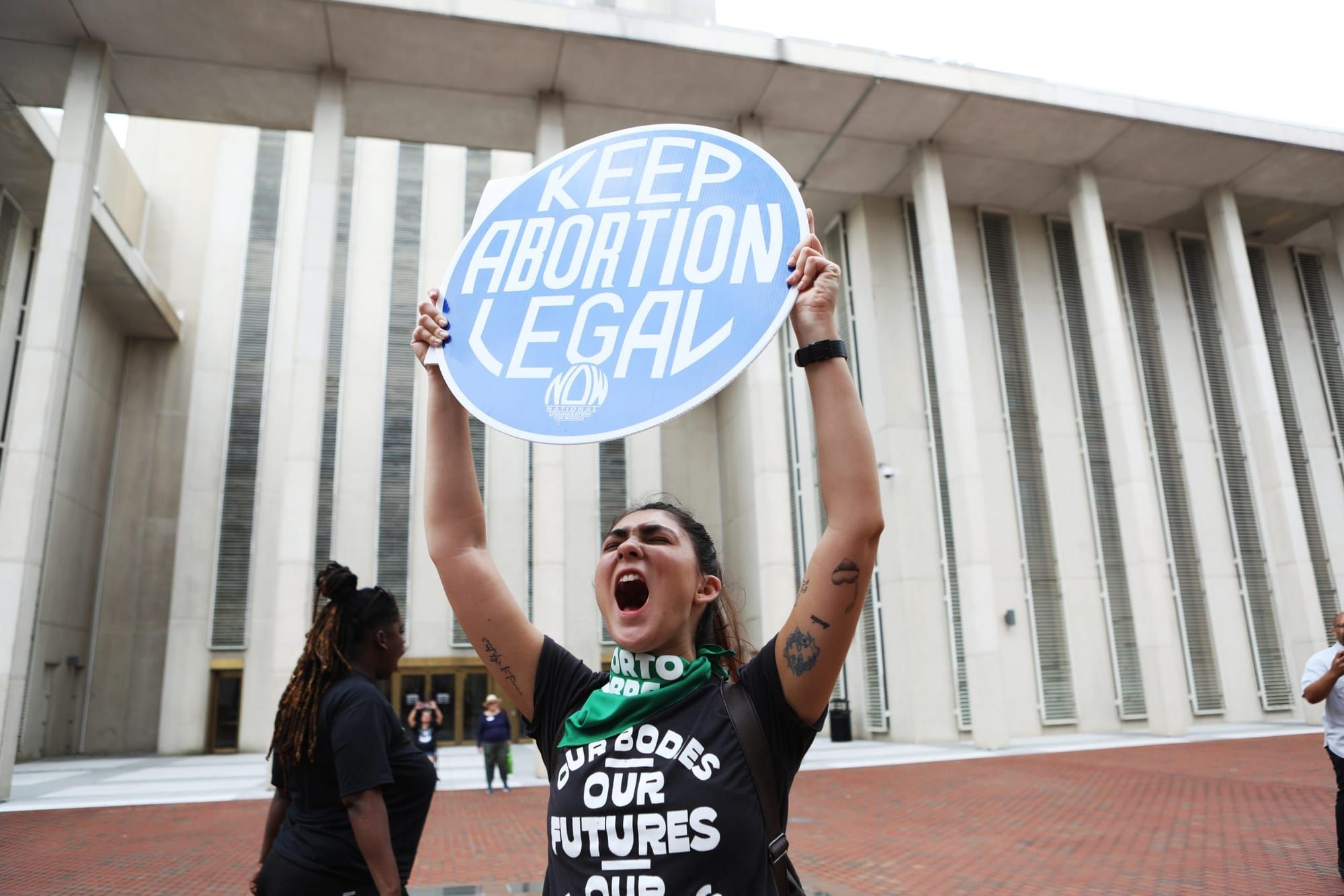 color photograph of an outdoor protest. a woman wearing a black t-shirt and green bandana holds a circular blue sign that rea