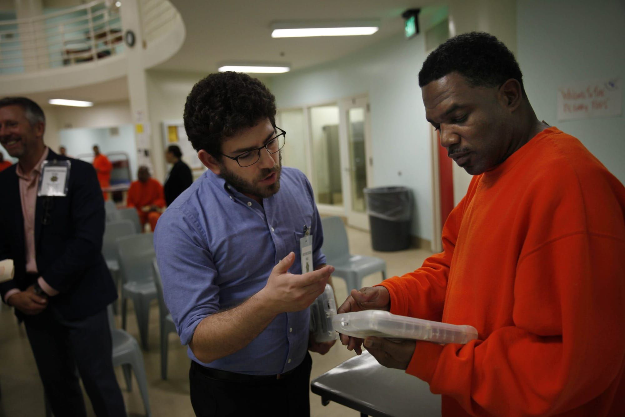 color photograph inside a prison. a black man in an orange sweater holds a tablet while a man in a blue button-down stands to