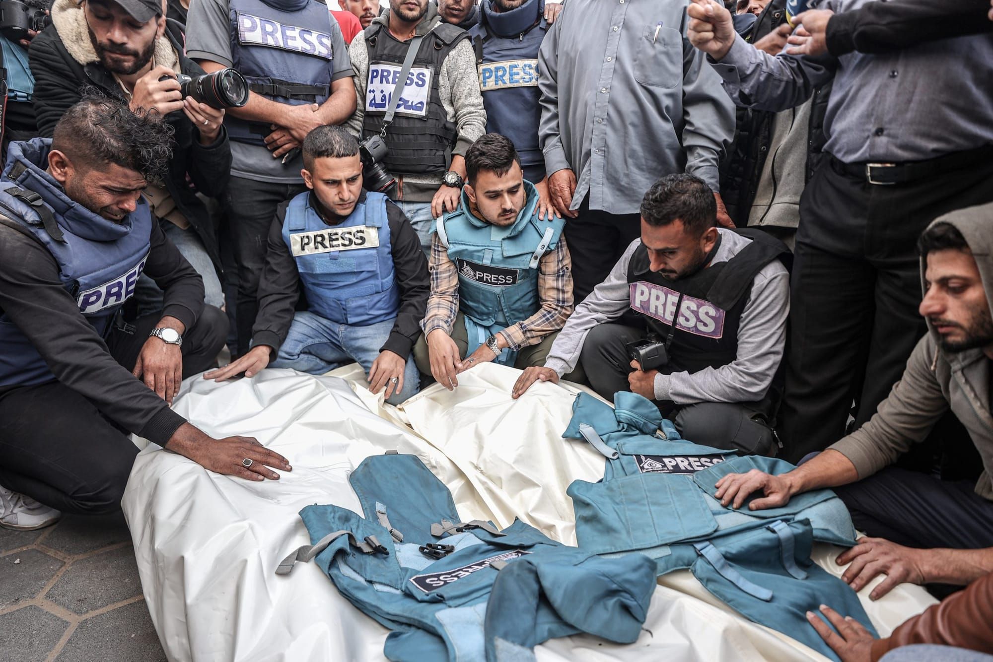 color photograph of people wearing blue and grey press vests kneeling around bodies in white cloth shrouds