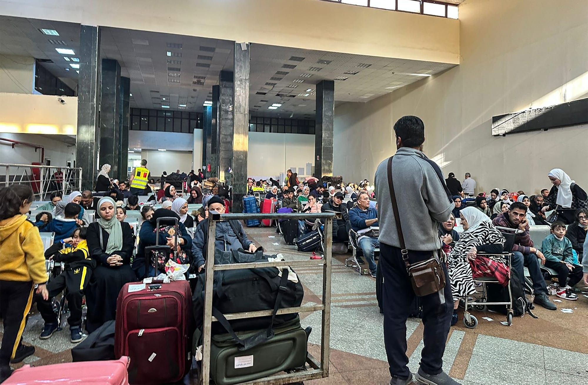 color photograph of people sitting in rows of chairs in a large room filled with luggage