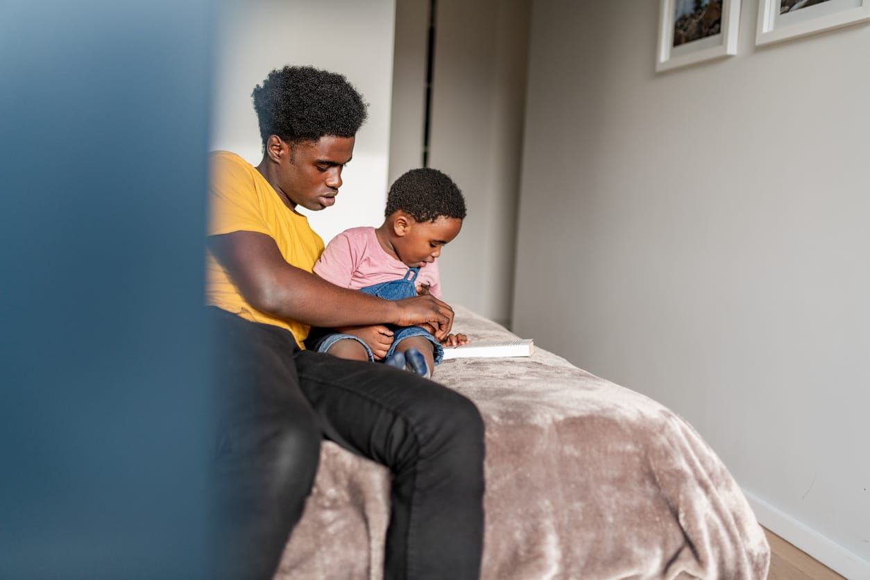 color photograph of a young Black father sitting on a bed holding his toddler while reading together