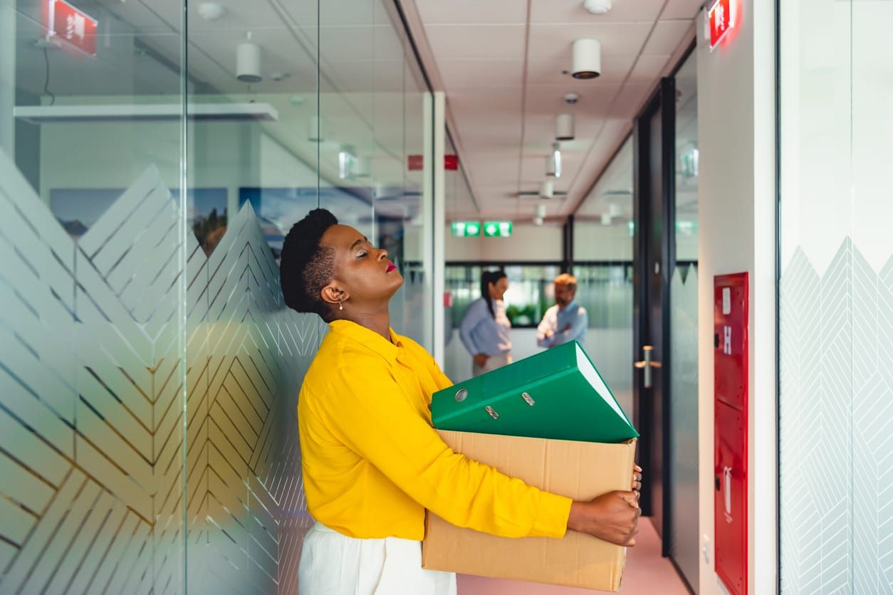 color photograph of a Black woman in a vibrant yellow shirt and white pants holding a cardboard box in the hallway of a corpo