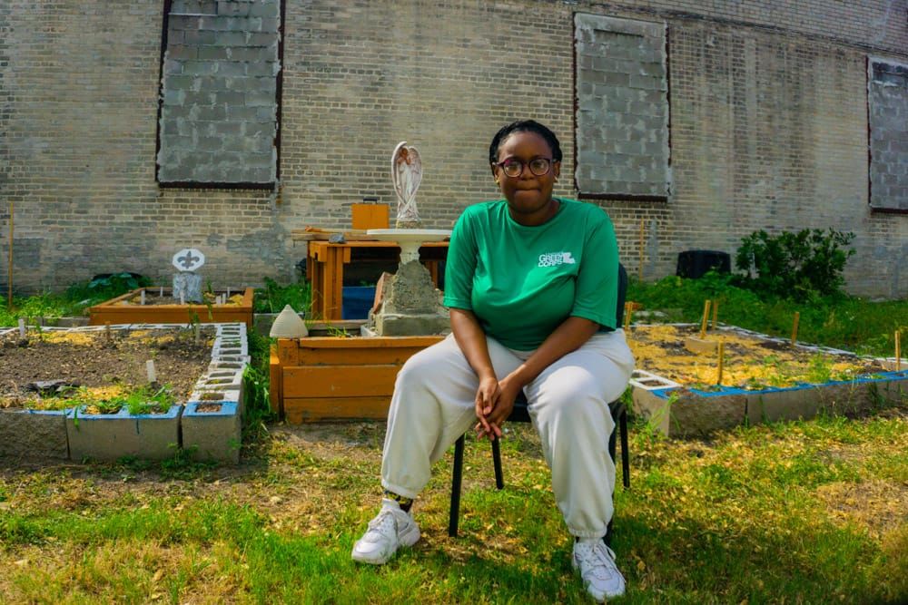 color photograph of a young Black woman with her hair tied back sitting in a chair in a grassy area with garden blocks. she w