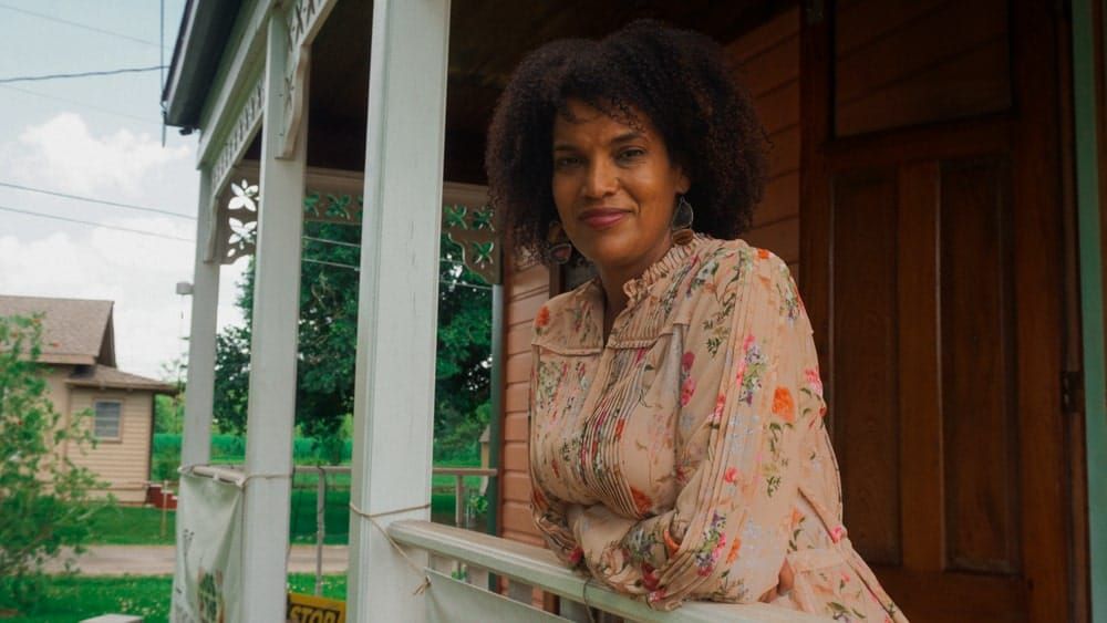 color photograph of a Black woman in a floral pink blouse leaning on the white wooden railing of a house