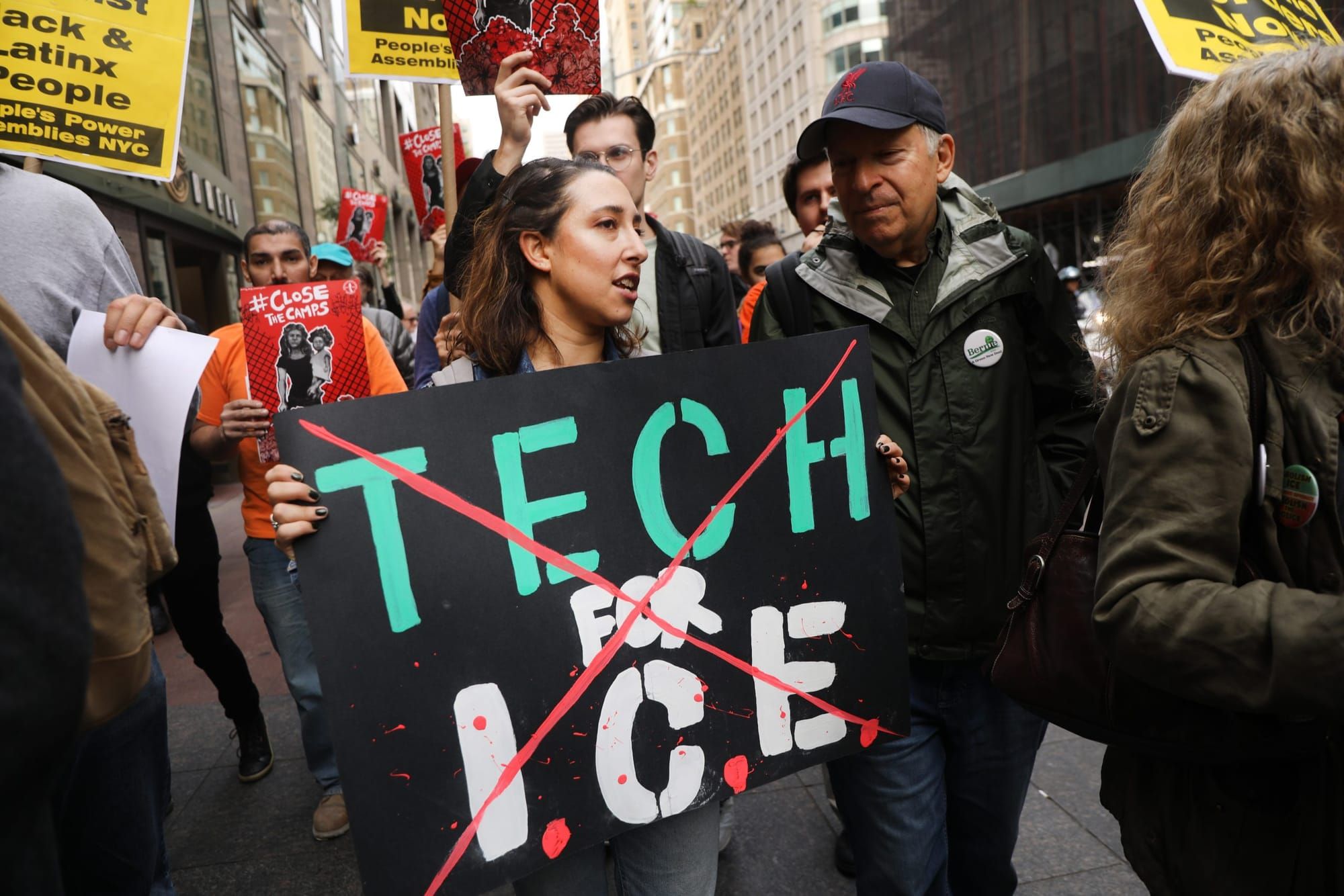 color photograph of an outdoor protest. a woman holds a large black poster with the words "tech for ICE" partially covered by
