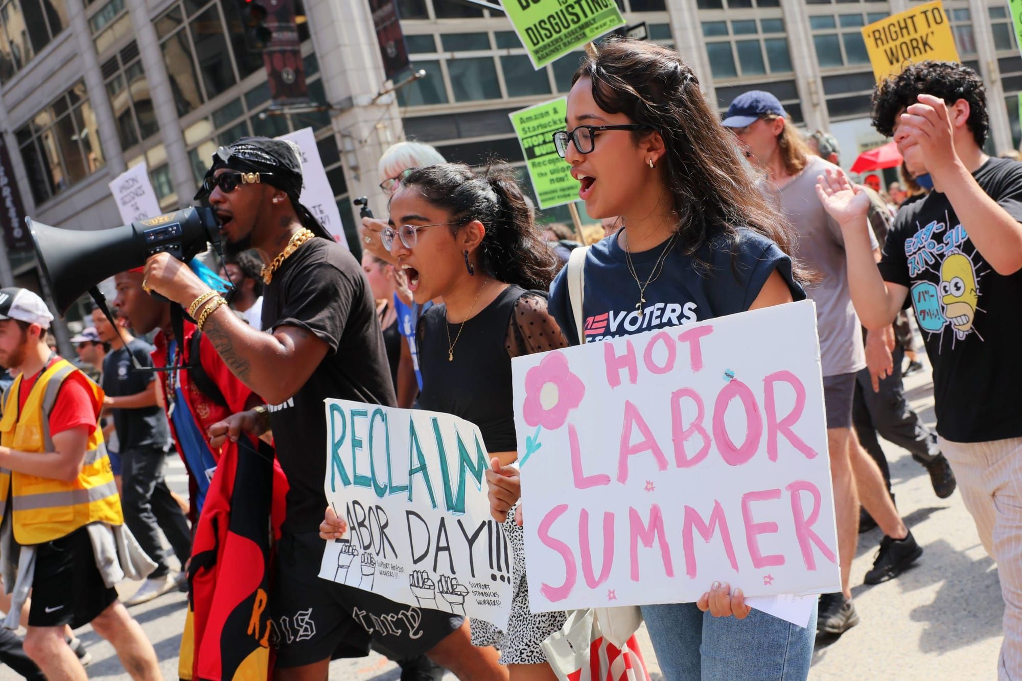 color photograph of an outdoor protest. two people walk by in the foreground holding posters with handwritten text reading "H