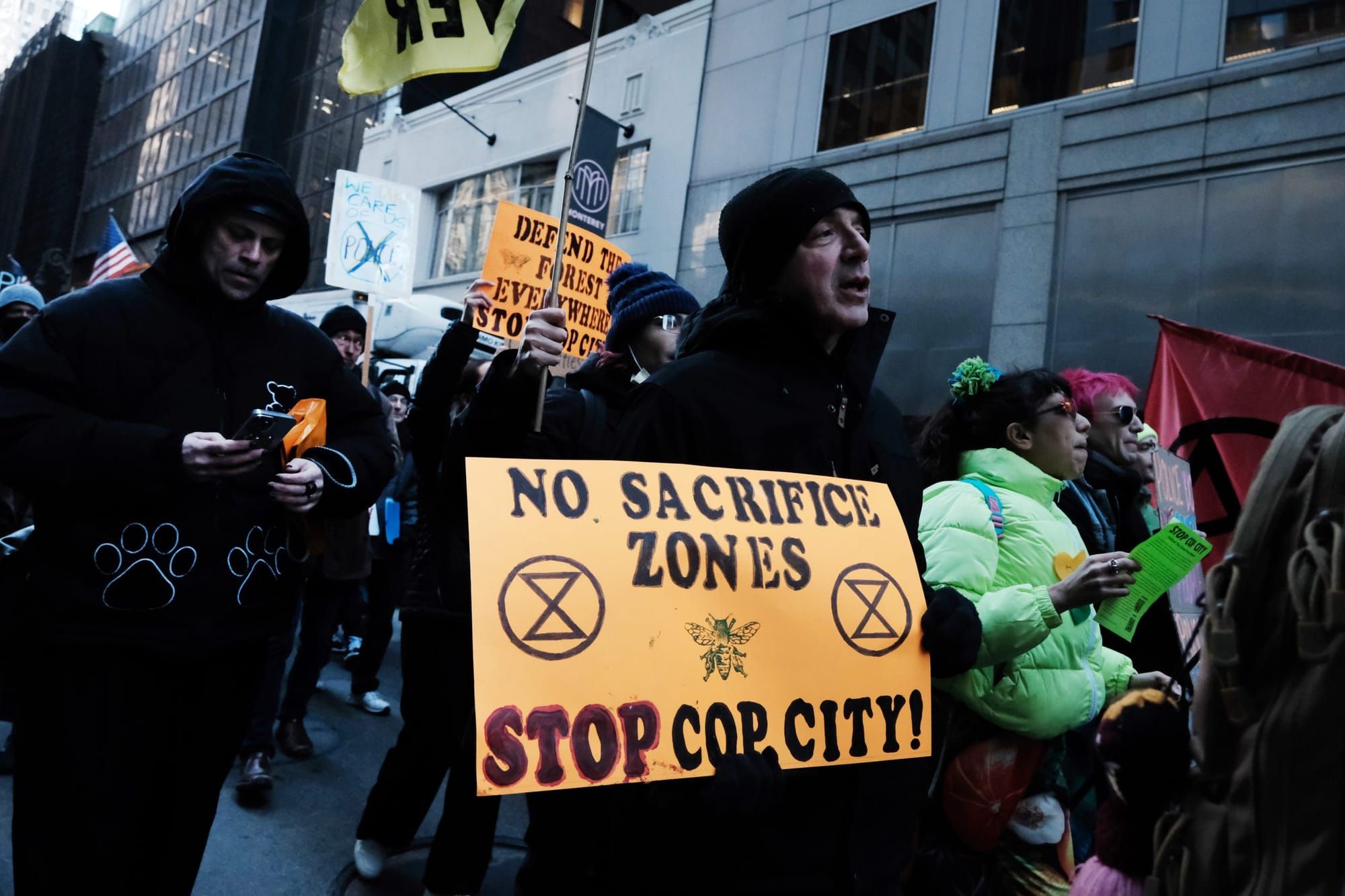 color photograph of an outdoor protest. people wearing dark colors walk in the middle of the street. one person in the center