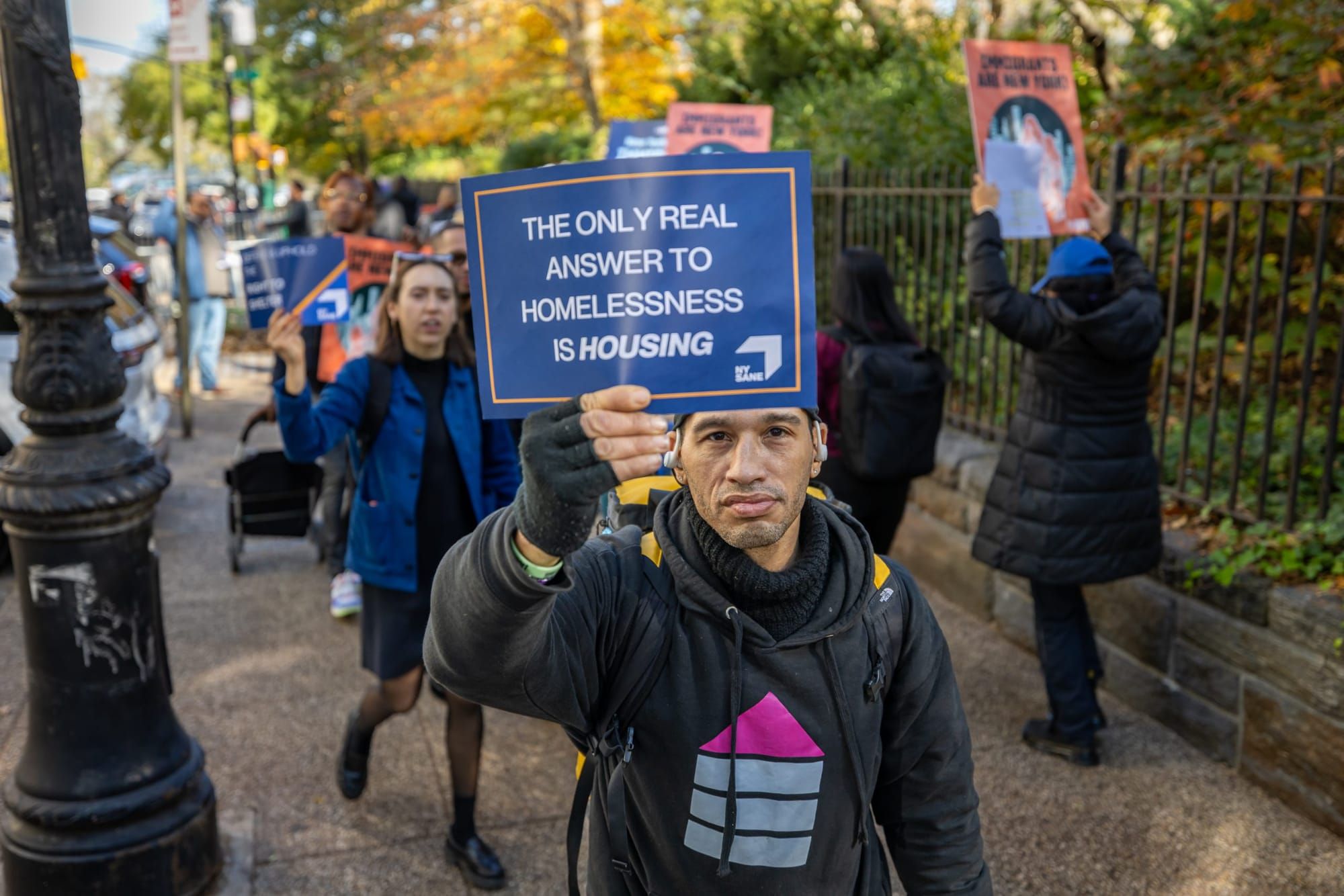 color photograph of an outdoor protest. people walk along a sidewalk, and a man in the foreground holds a blue poster with te