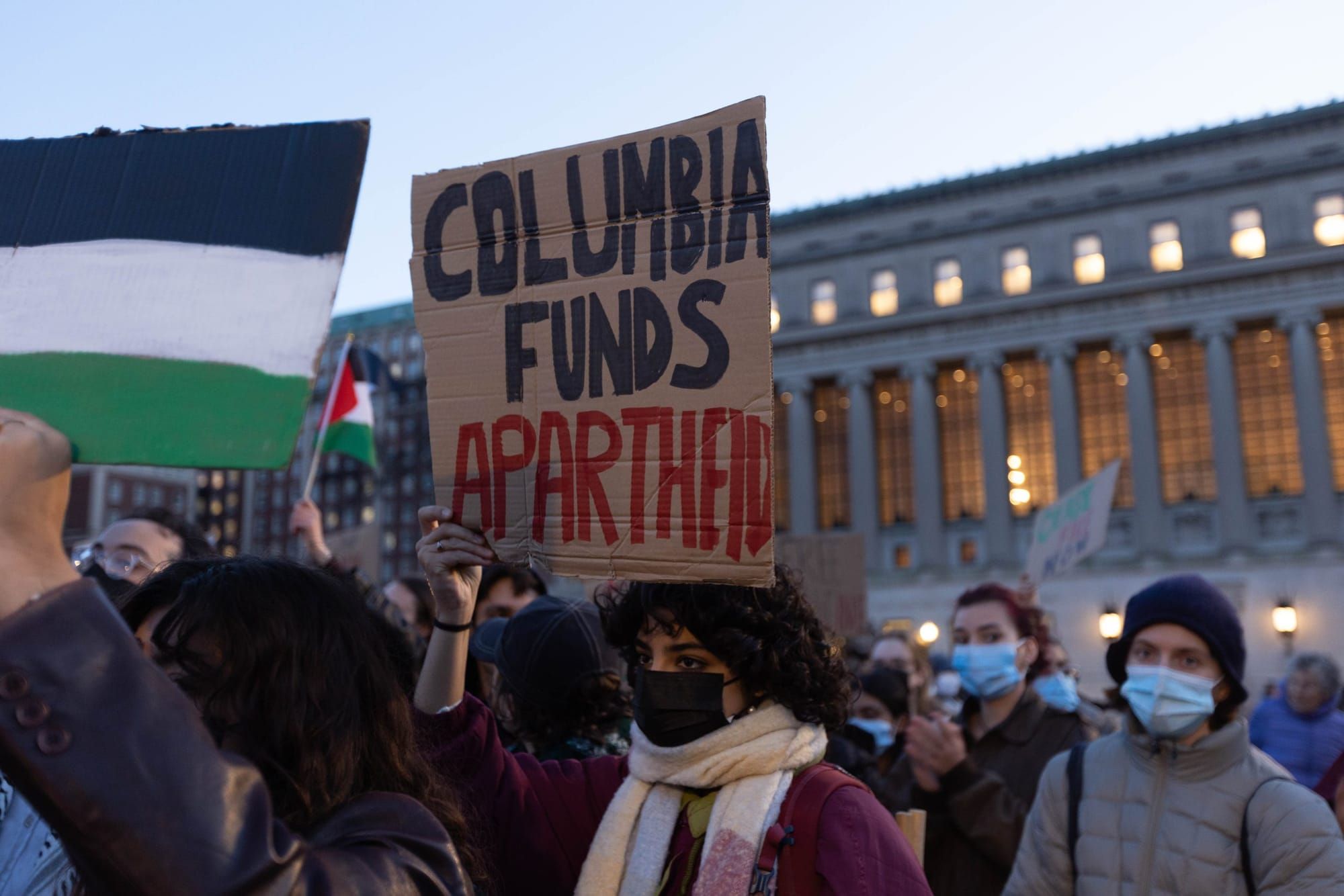 color photograph of an outdoor protest in support of palestine. in the center, someone holds a cardboard sign that reads "col