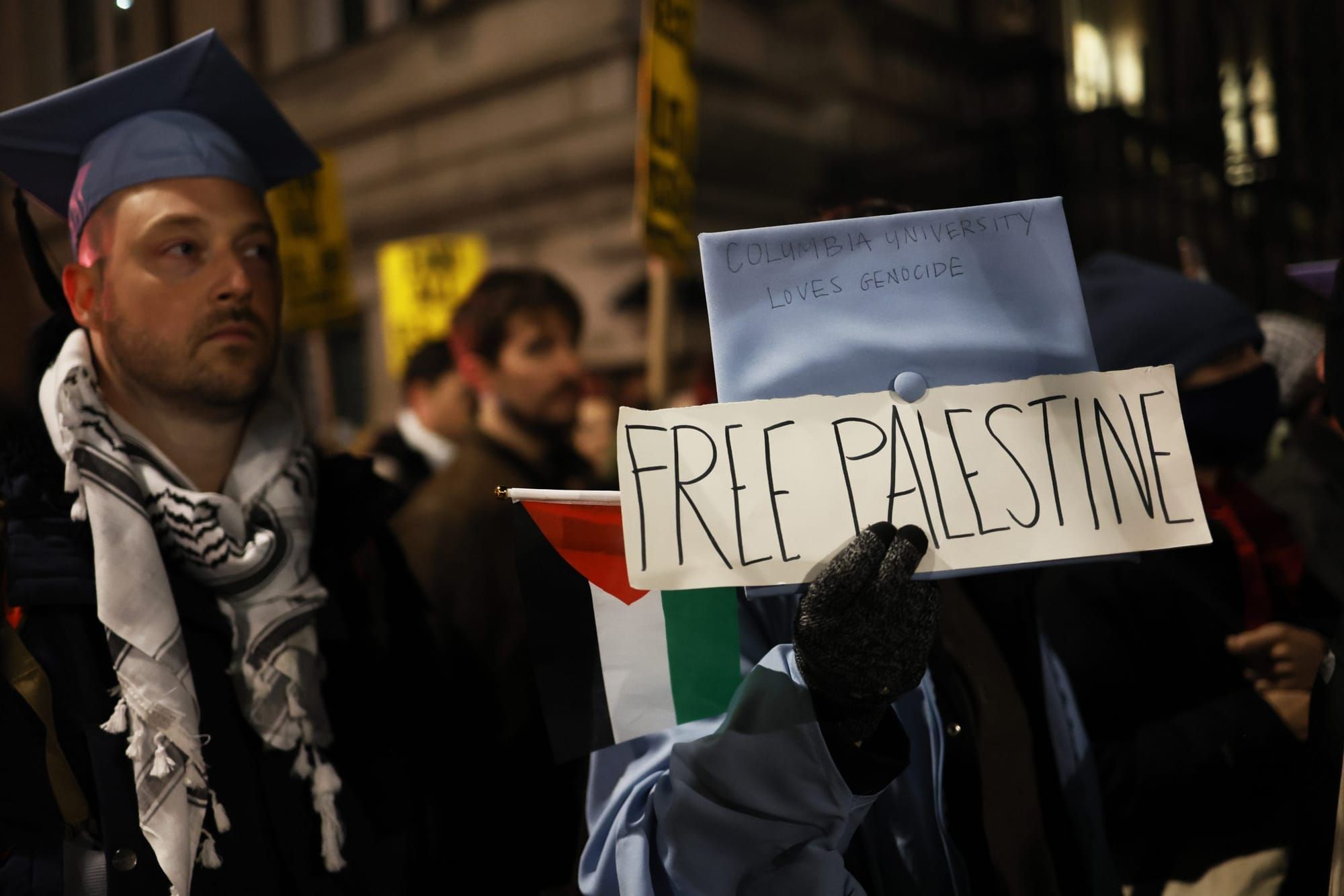 color photograph of a nighttime protest. people wear Columbia blue graduation caps and keffiyehs. one person holds a poster i