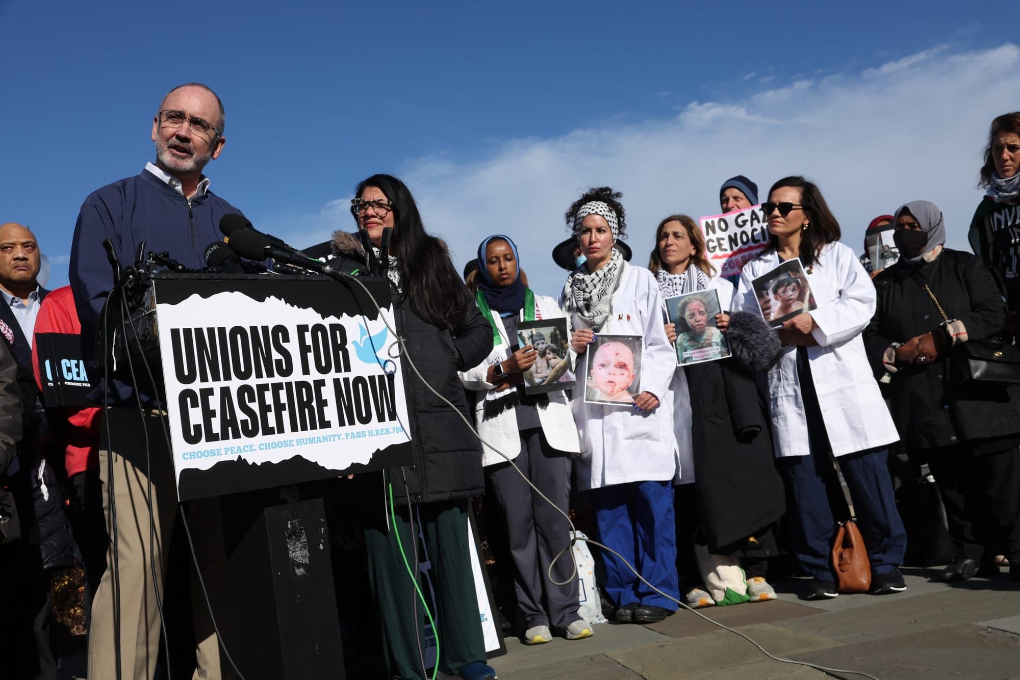 color photograph of an outdoor press conference. shawn fain stands in a black sweater at a podium; a poster on the podium rea