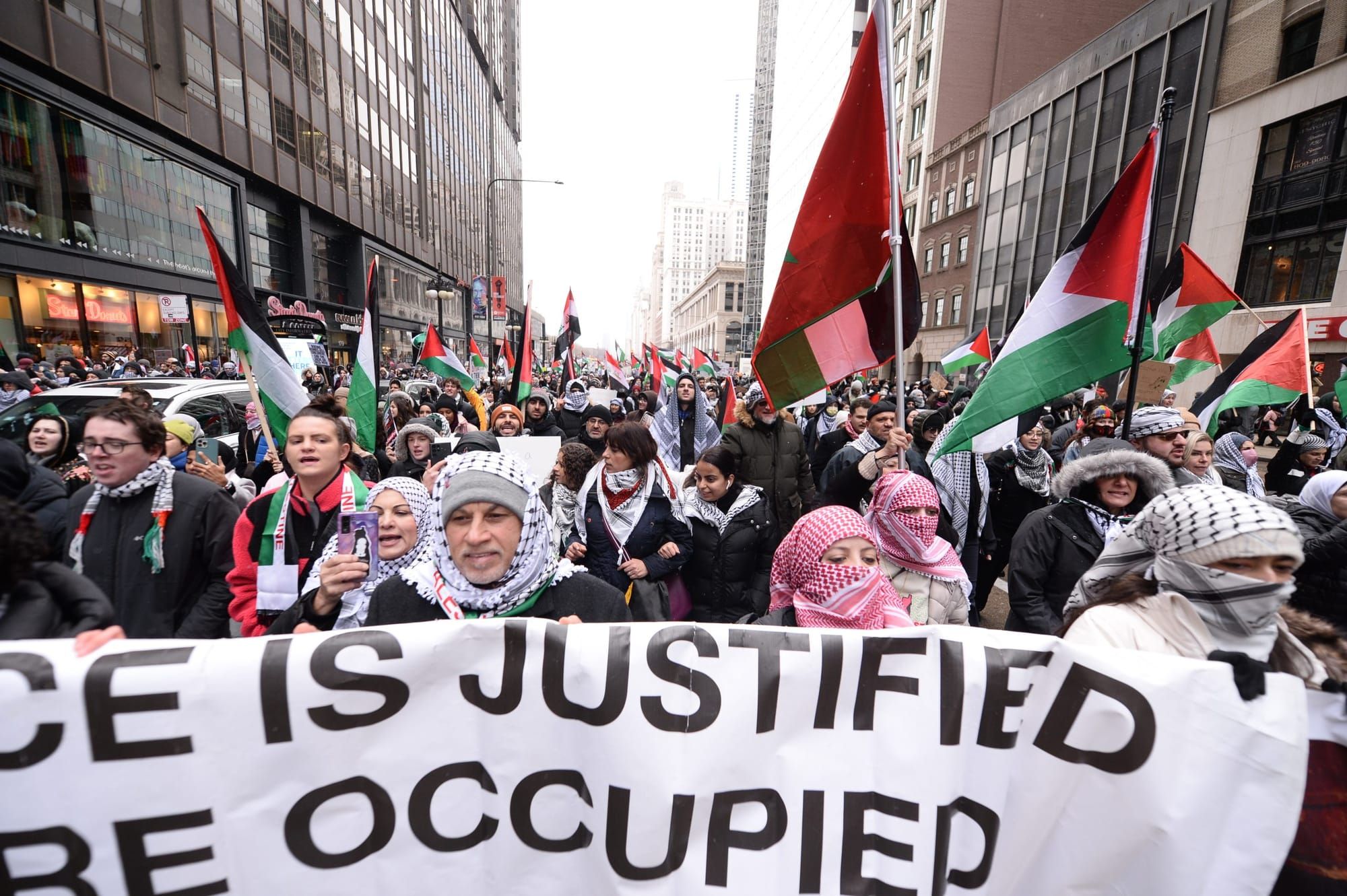 color photograph of an outdoor protest in support of palestine. people wearing keffiyeh hold a large white banner with a pale
