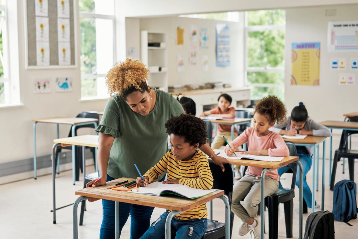 color stock photo of a Black woman teacher standing over a desk in her classroom to help her elementary school student