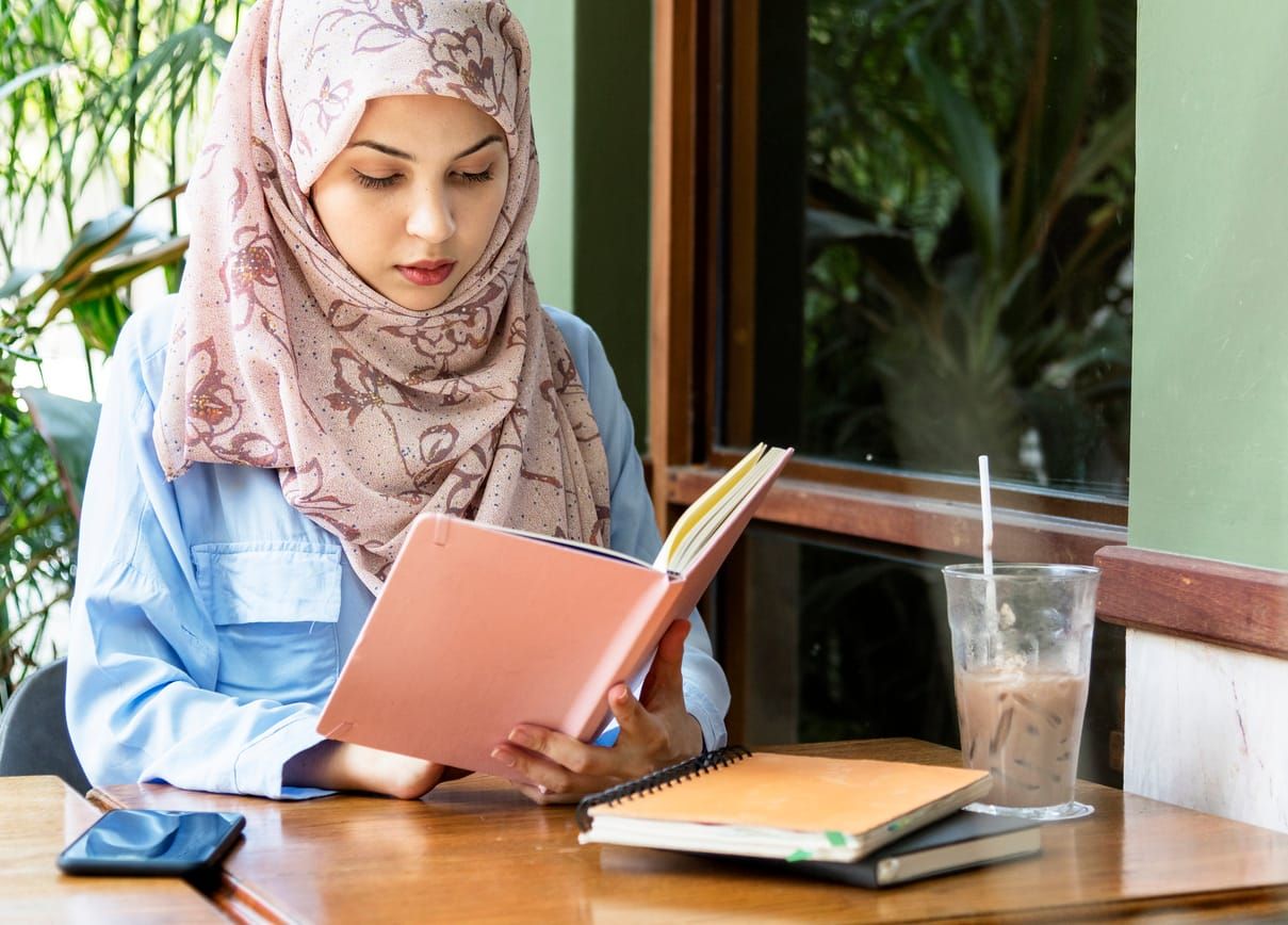 Brown woman wearing a pink head scarf reading book at a cafe table