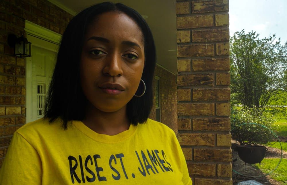 color photograph of a Black woman in a yellow t-shirt with black text reading "Rise St. James." she stands in front of the co