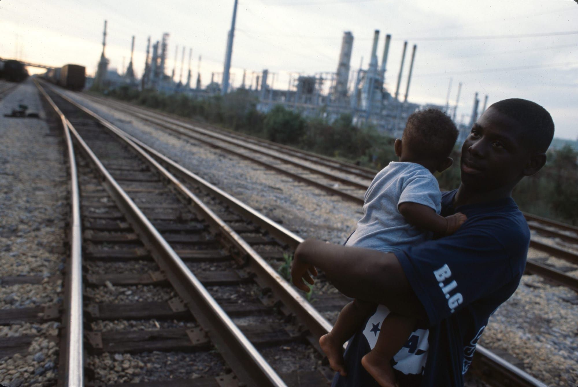 color photograph of a Black man holding his baby while standing on railroad tracks leading to an oil refinery plant