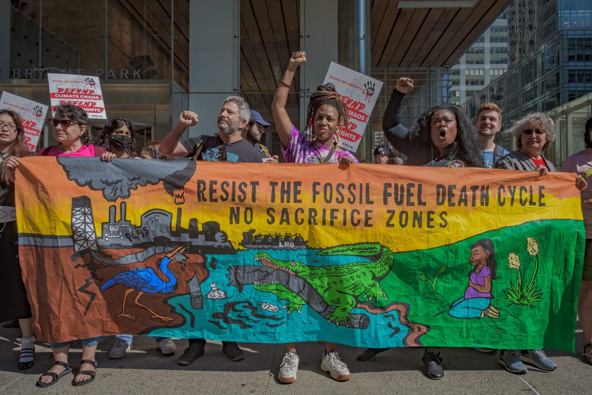 color photograph of an outdoor climate protest. people hold a large multi-colored banner with black text reading "resist the