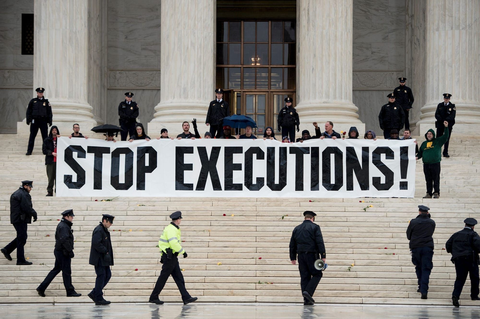 color photograph of an outdoor protest on a government building's steps. people hold a large white banner with black text tha