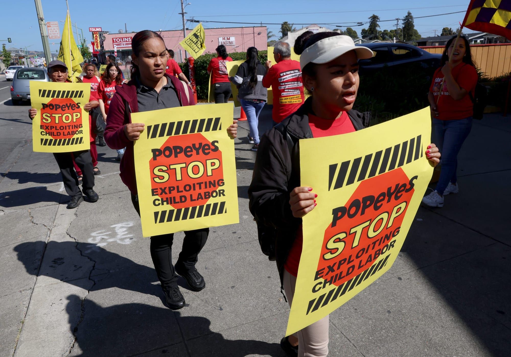 color photograph of an outdoor protest against child labor. people walk in a line towards the camera holding yellow signs rea