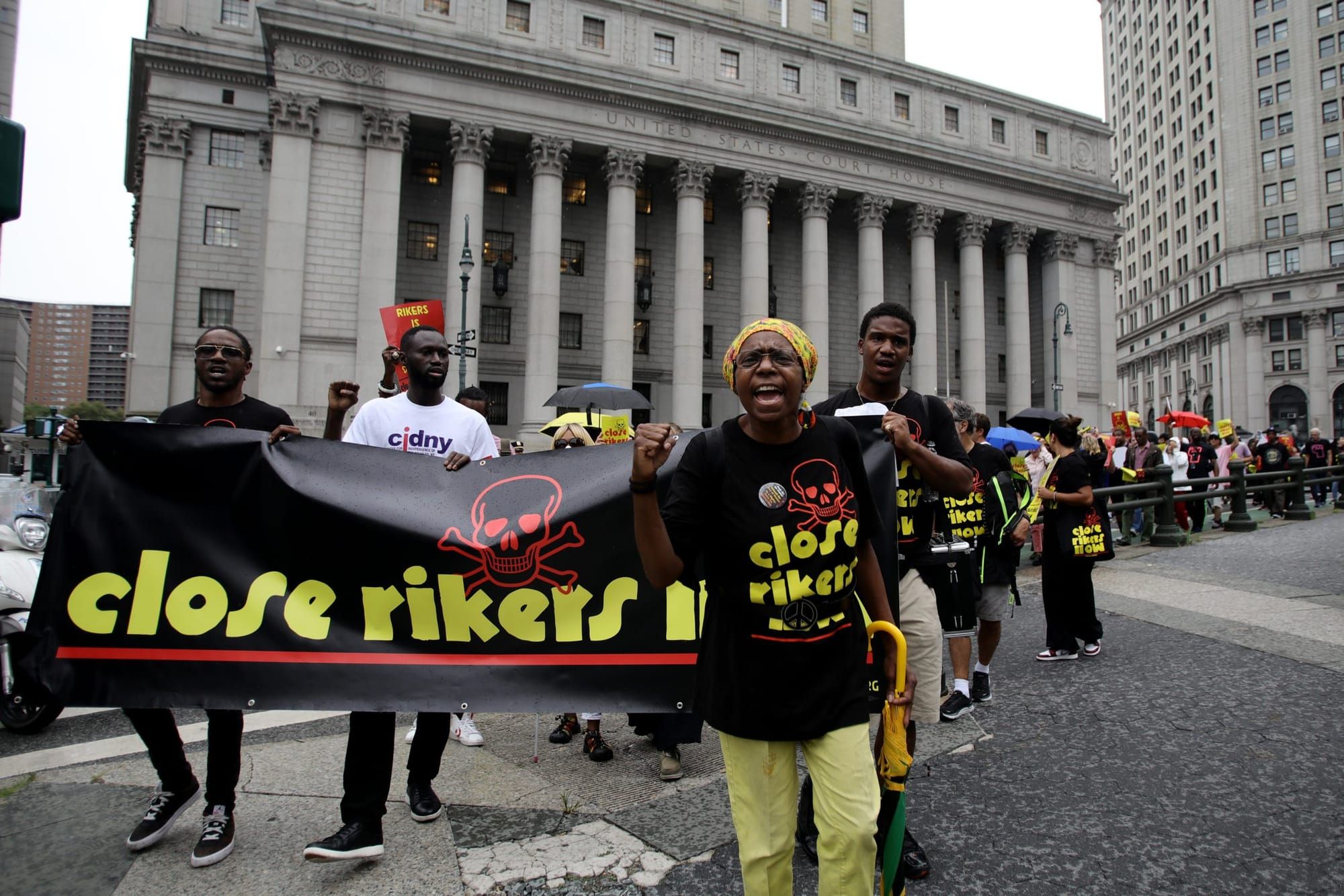 color photograph in protest of Rikers Island. people hold a black banner with bright yellow text that reads "close rikers"