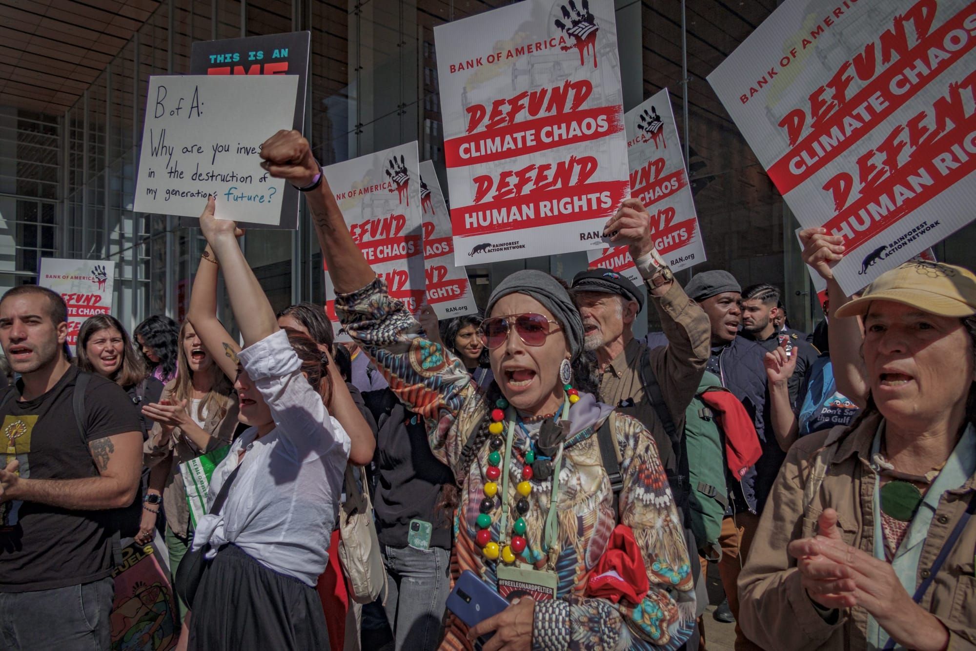 color photograph of an outdoor protest in favor of climate justice. people hold up white posters with red text reading "bank