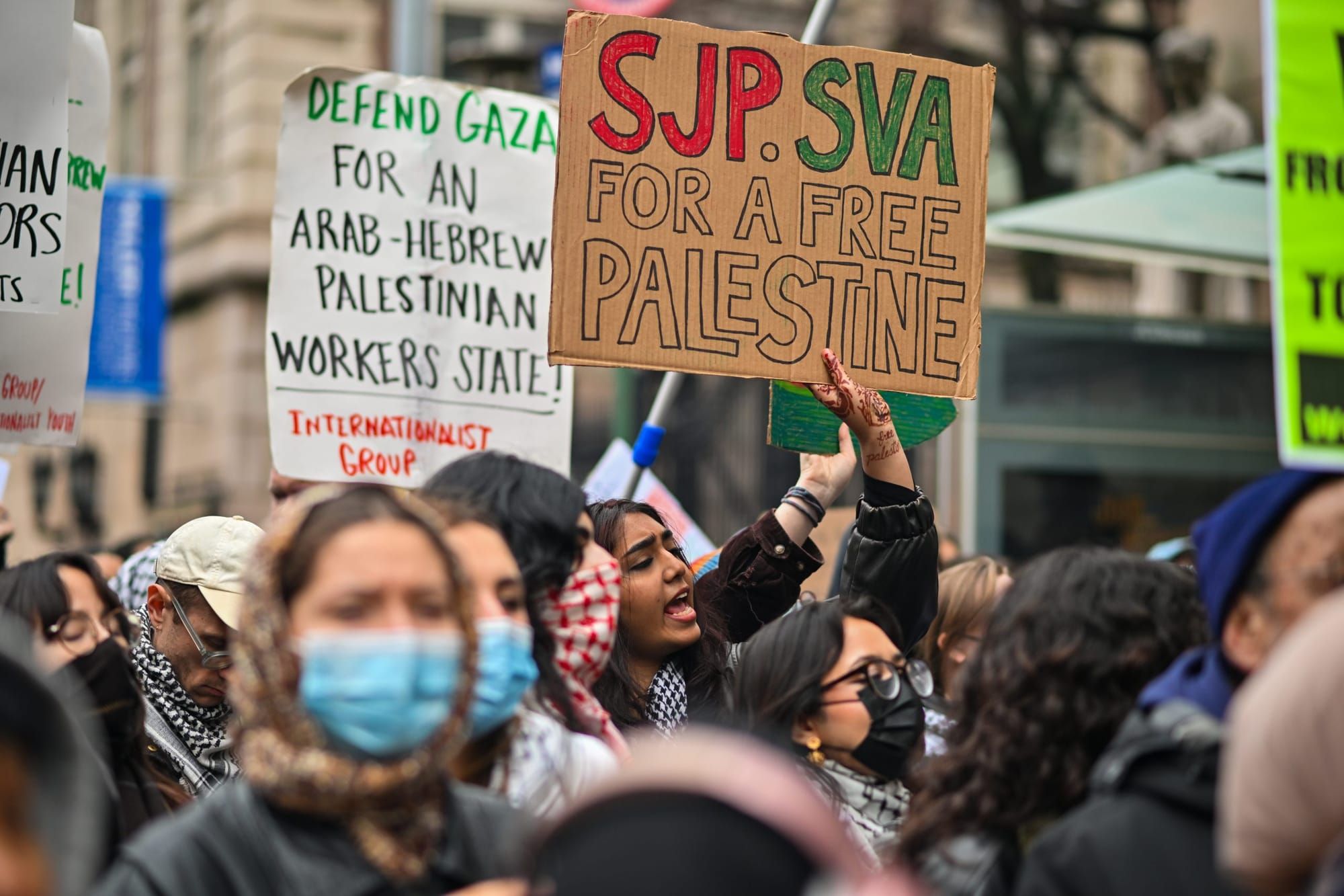 color photograph of an outdoor protest in support of Palestinian liberation. people wear face masks as they hold signs readin