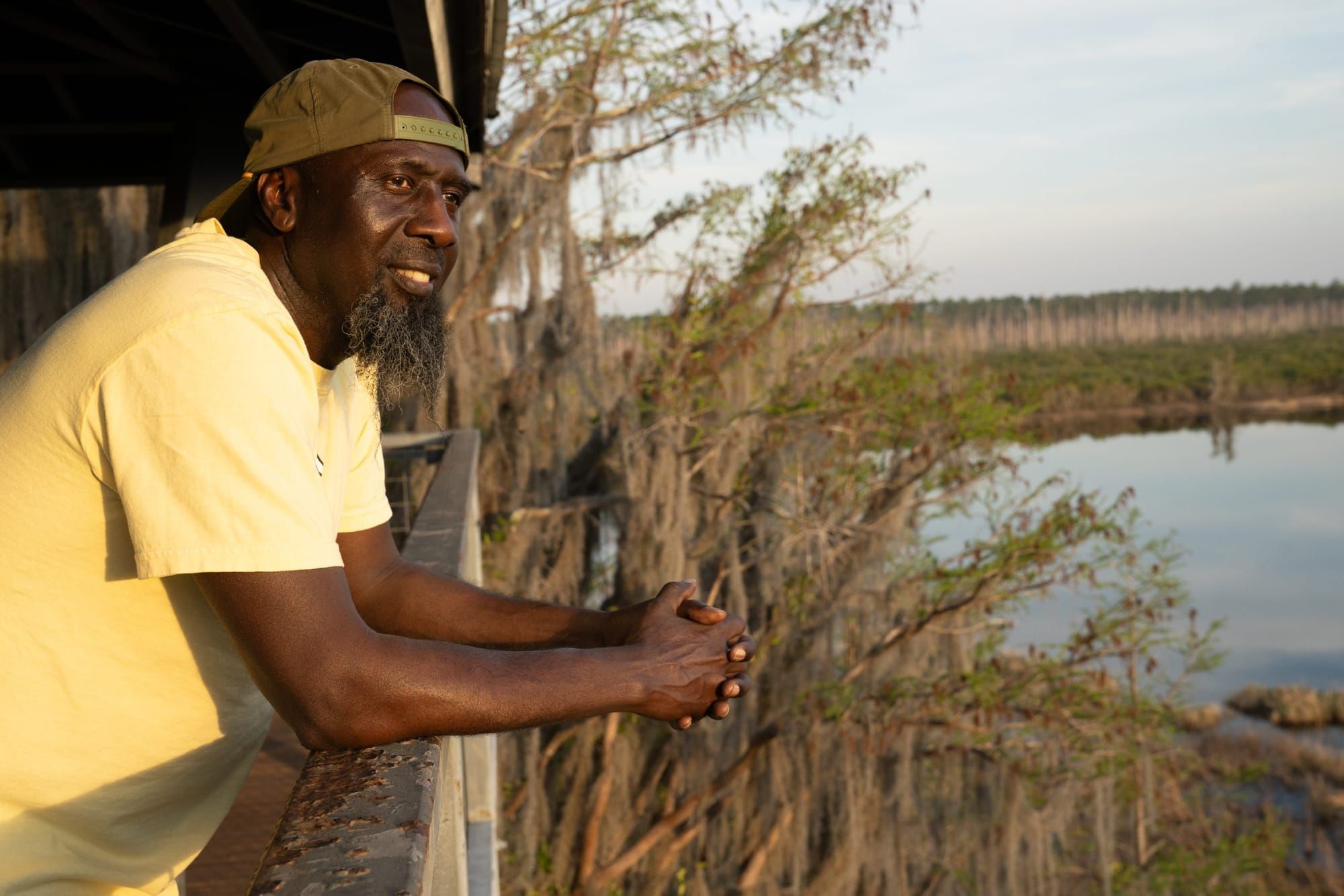 color photograph of a Black man in a light yellow t-shirt leaning on a wooden balcony overlooking a swamp