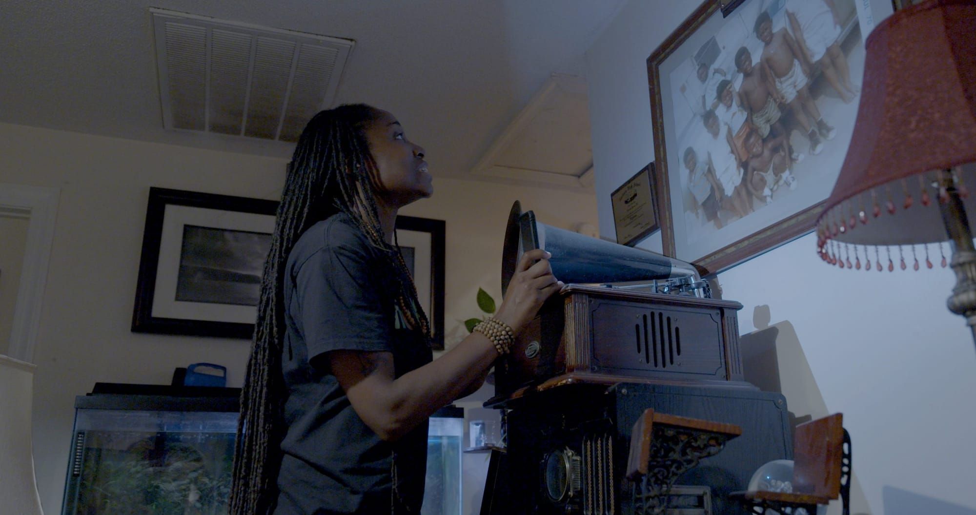 color photograph of a young Black woman in a dark room of a family house, staring at a photograph of young Black boys hanging