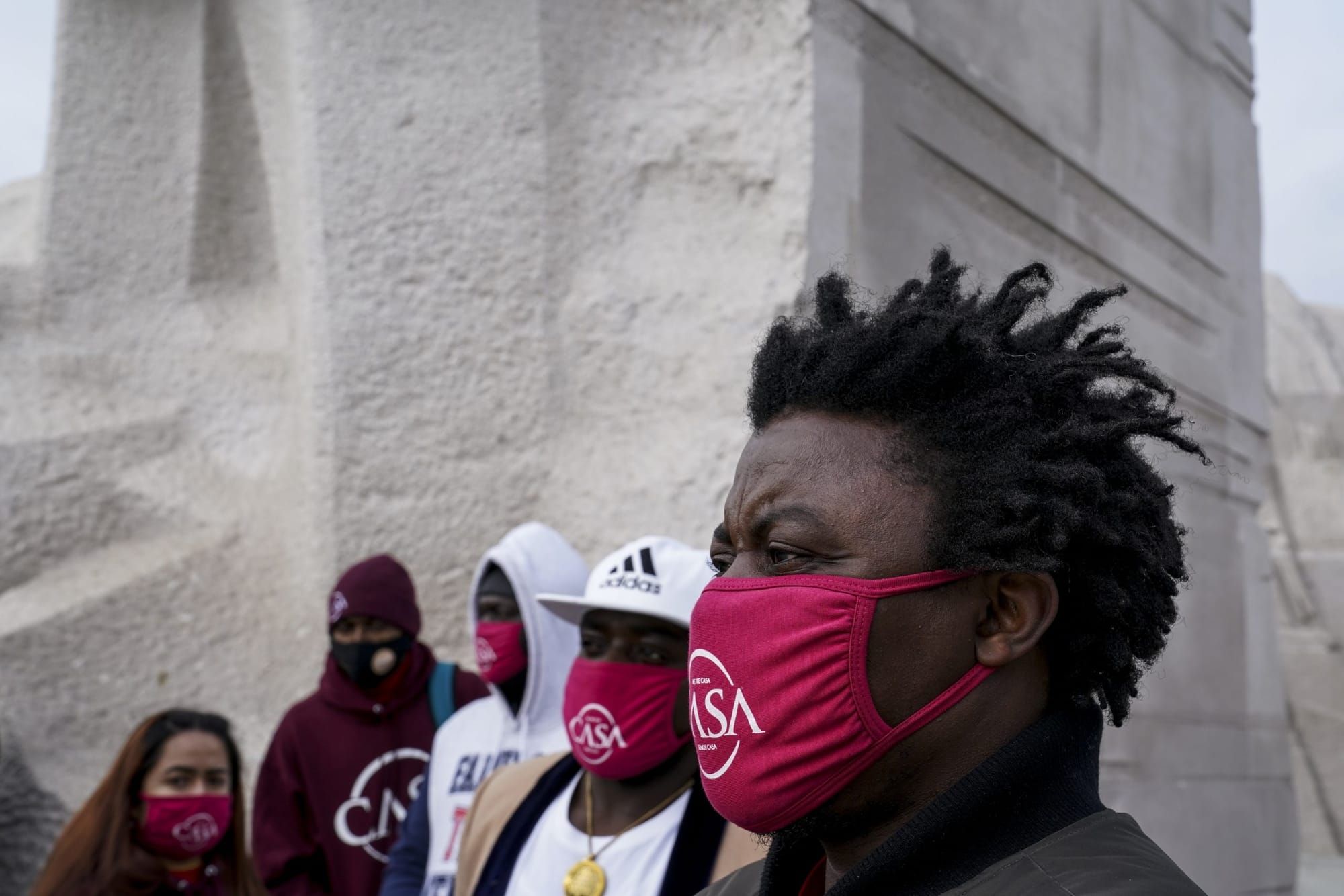 color photograph of Black people wearing red cloth face masks in front of a white stone statue
