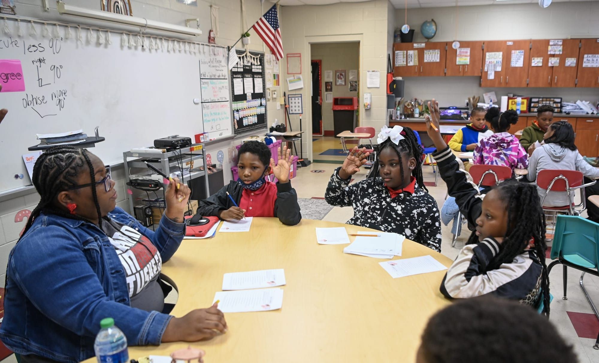 color photograph of a Black teacher sitting at a round table in a classroom with young elementary school-age children