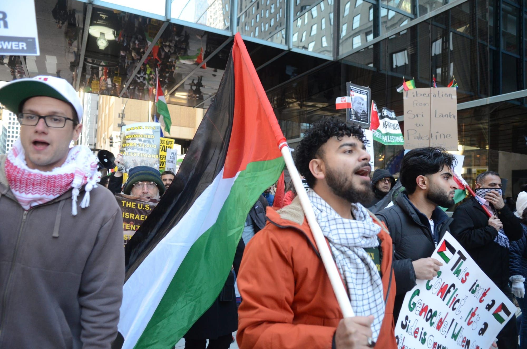color photograph of an outdoor protest in support of Palestinian liberation. a person wears a keffiyeh around his neck and ca