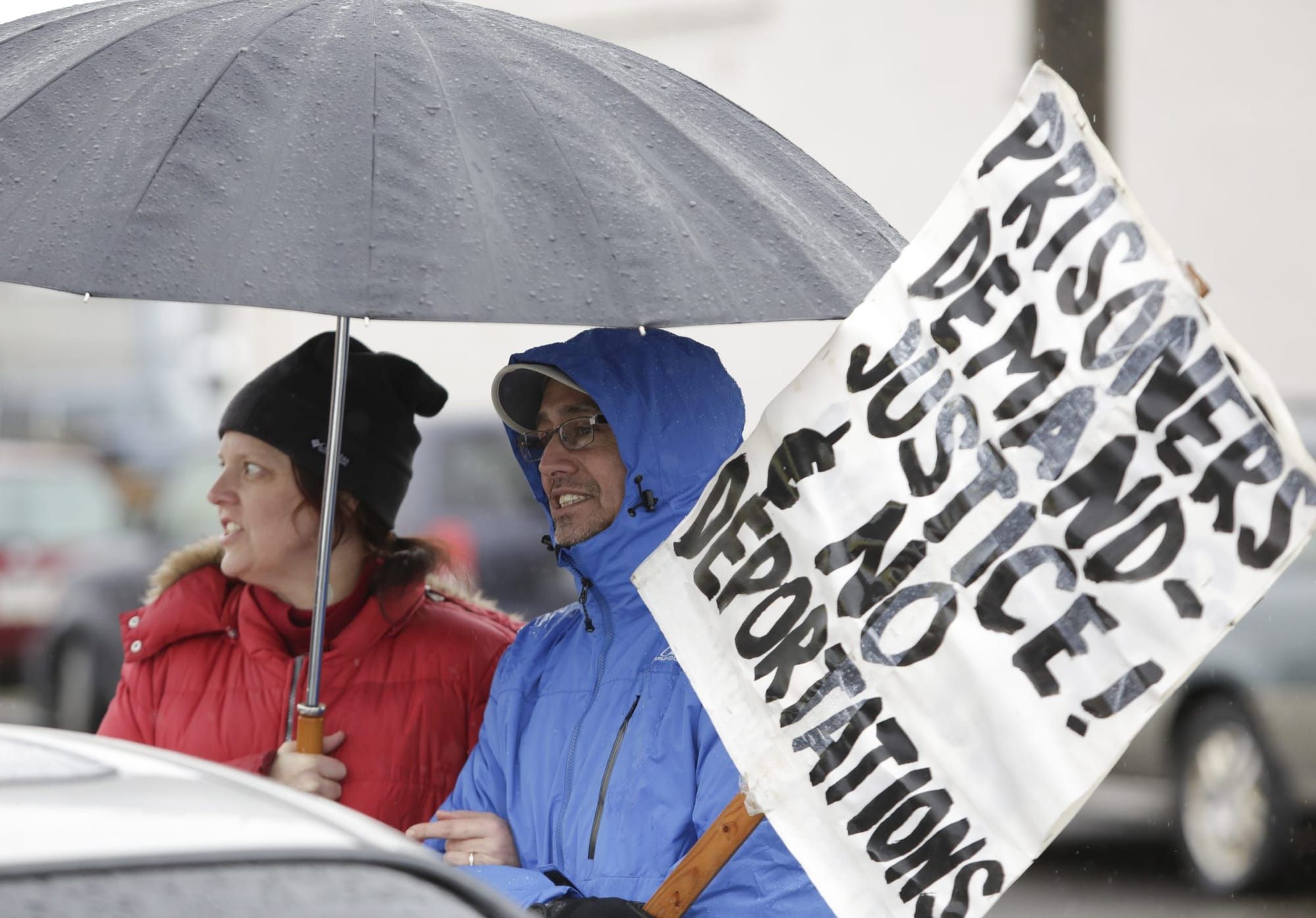 color photograph of an outdoor protest. two people stand under an umbrella; one holds a white picket poster that reads "priso