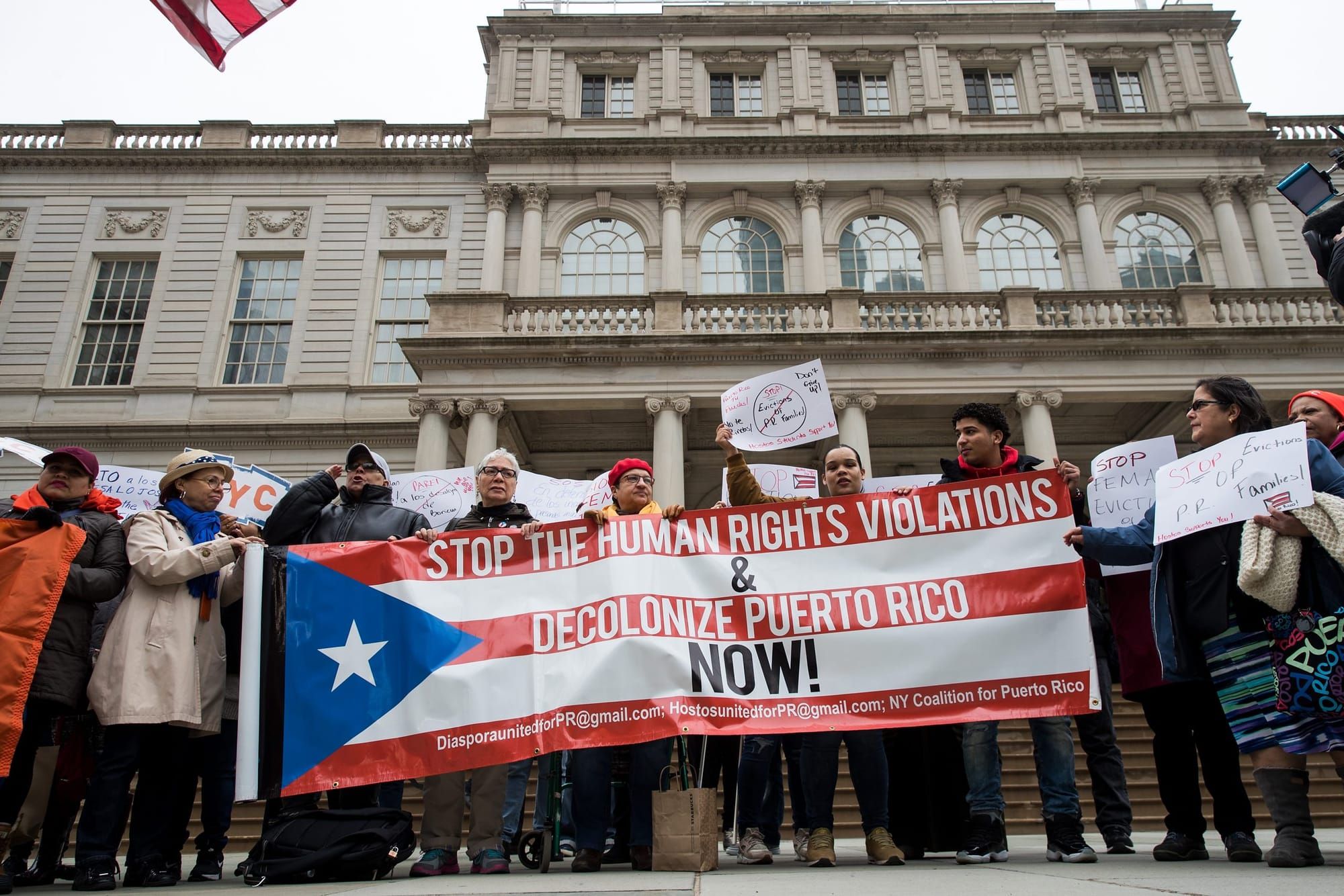 color photograph of an outdoor protest on the steps of a government building. people hold up a poster of a puerto rican flag